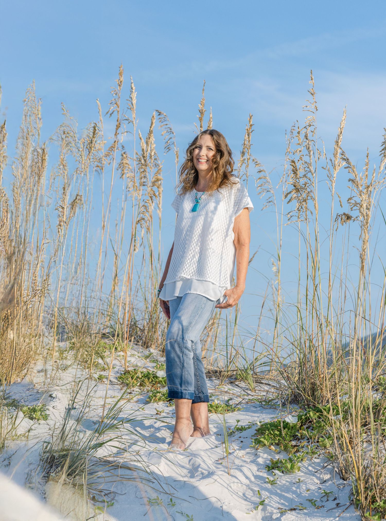 Woman in white top and denim skirt walks along sandy beach path with tall sea grass under blue sky.
