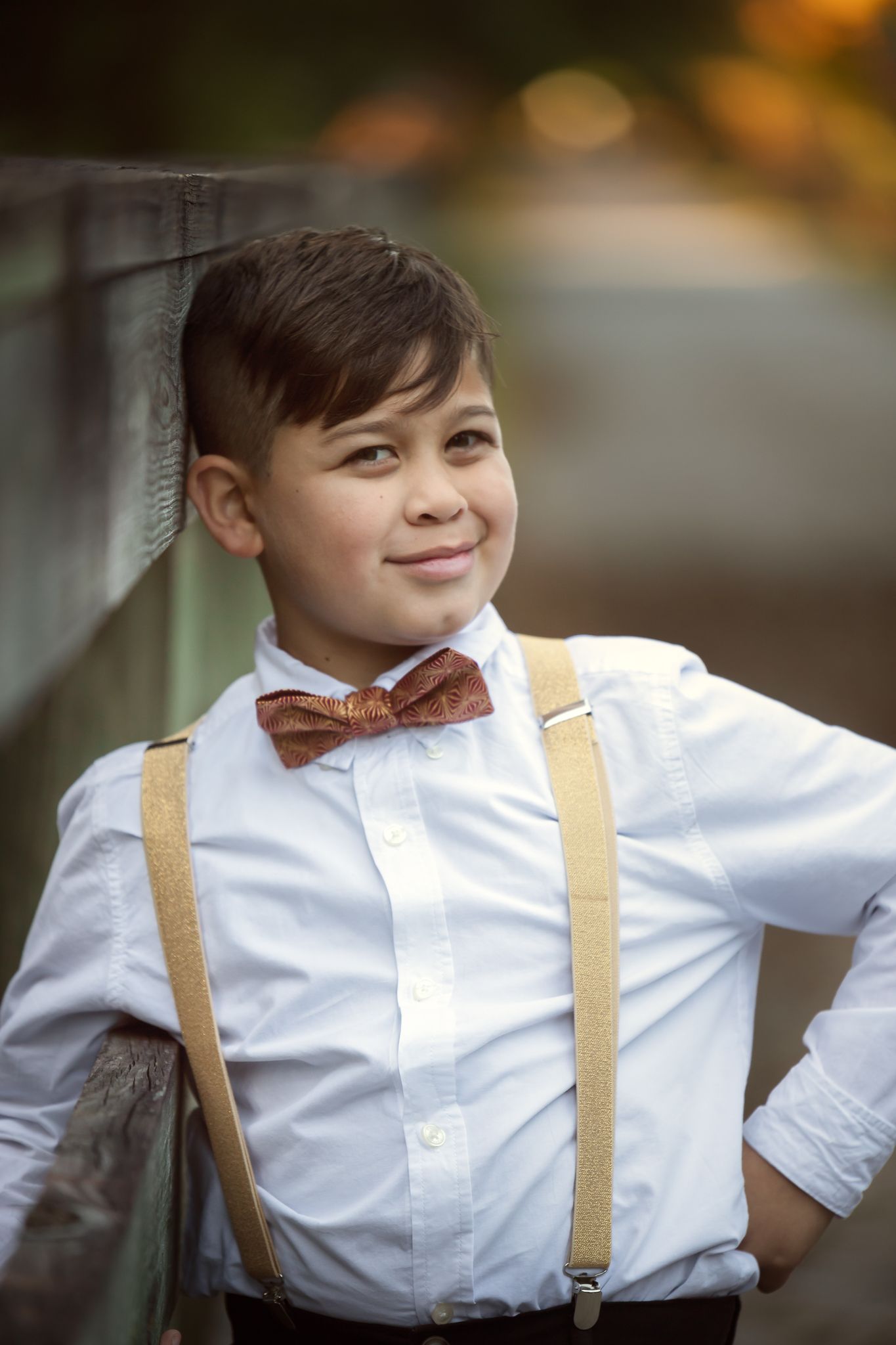 A young person in a formal outfit with a white dress shirt, brown bow tie and tan suspenders poses outdoors.