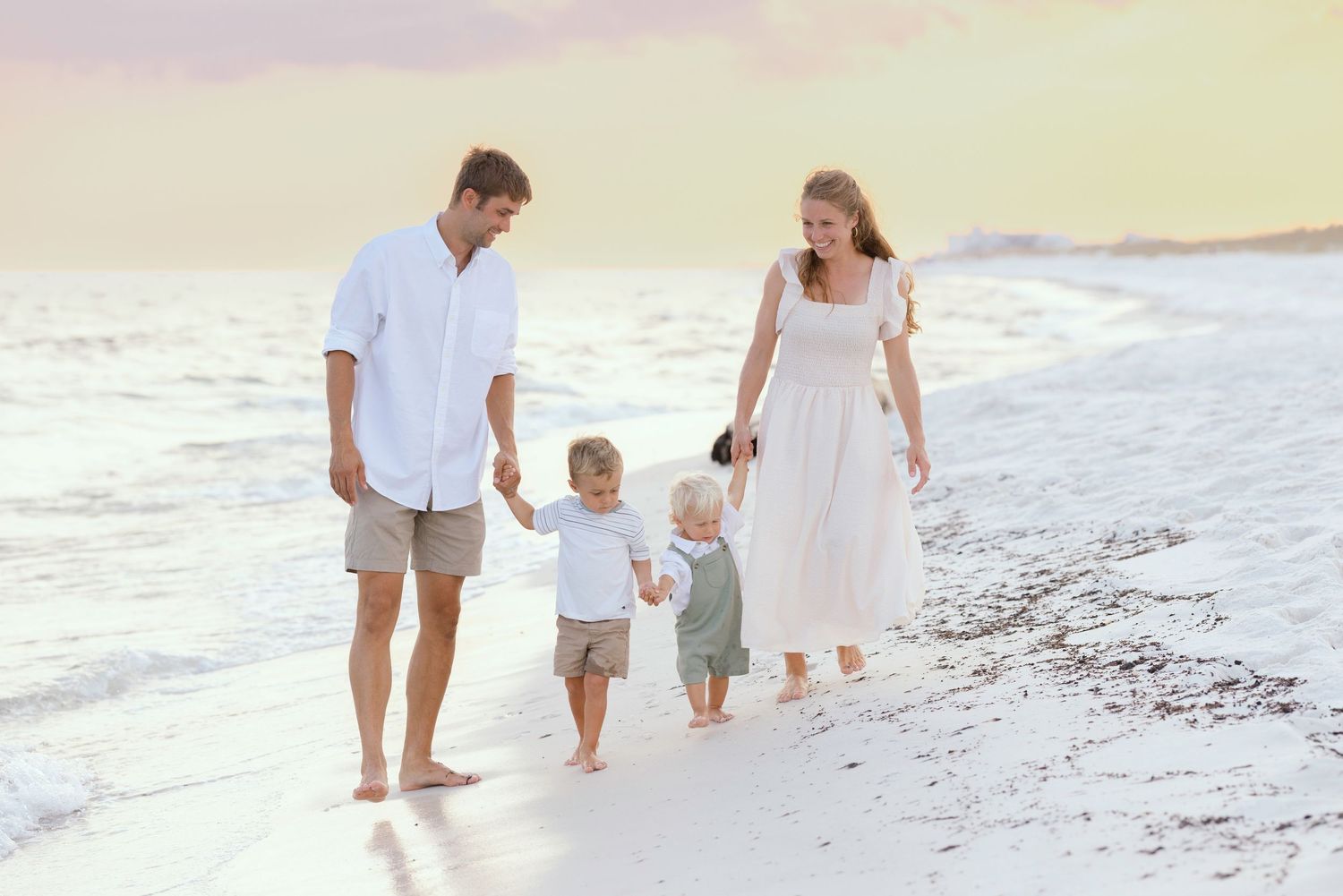 Family holding hands while walking along scenic beach shoreline at sunset in light colored casual attire.
