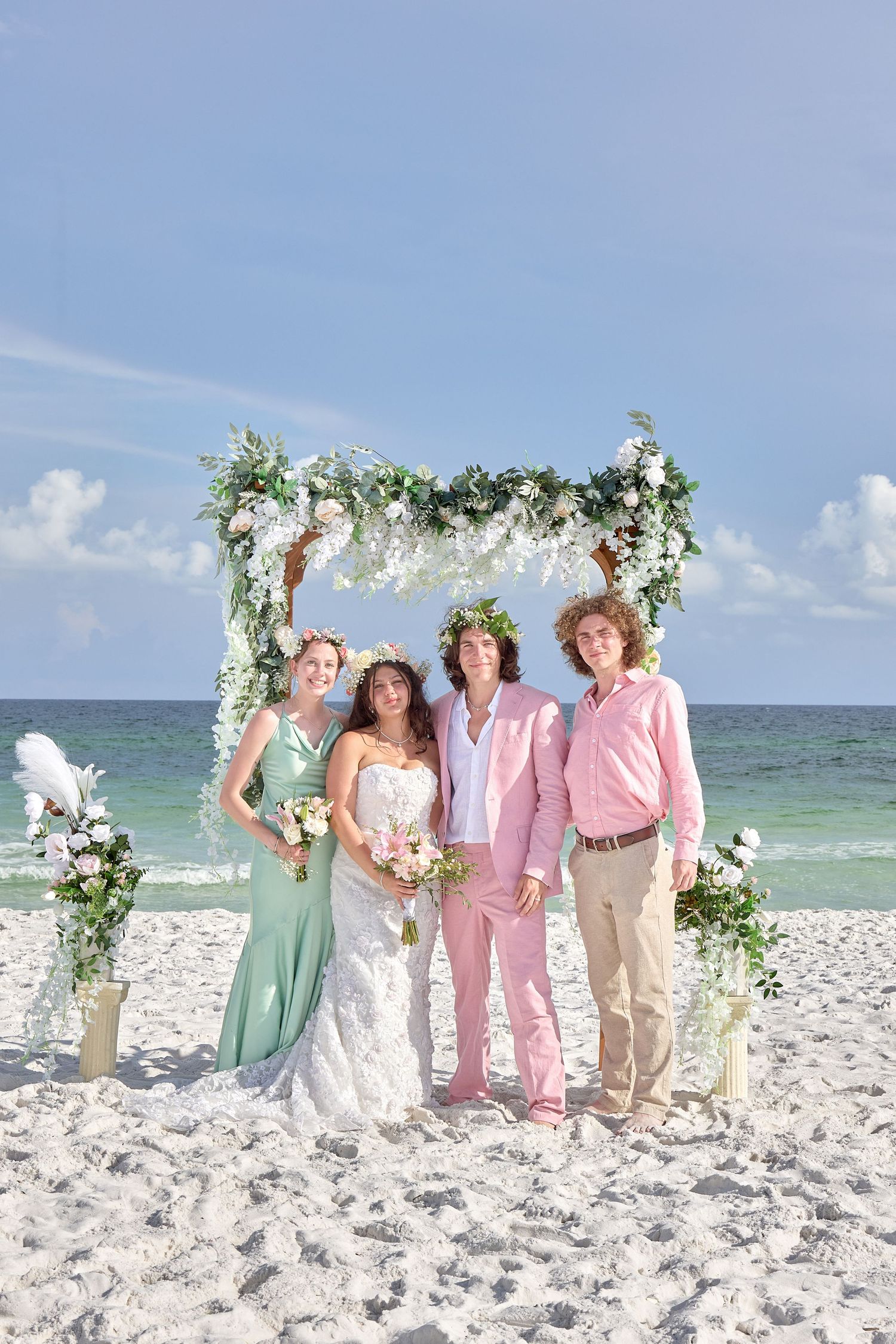 Beach wedding party poses under floral arch with ocean backdrop, bride in white, groom in pink suit, bridesmaid in mint green.