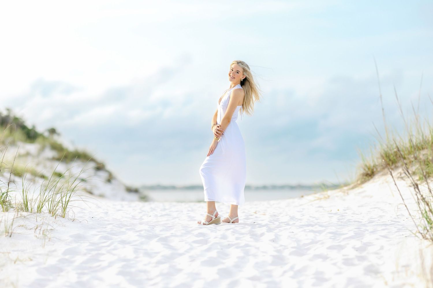 Woman in white dress standing on sandy beach with dunes and cloudy sky backdrop.