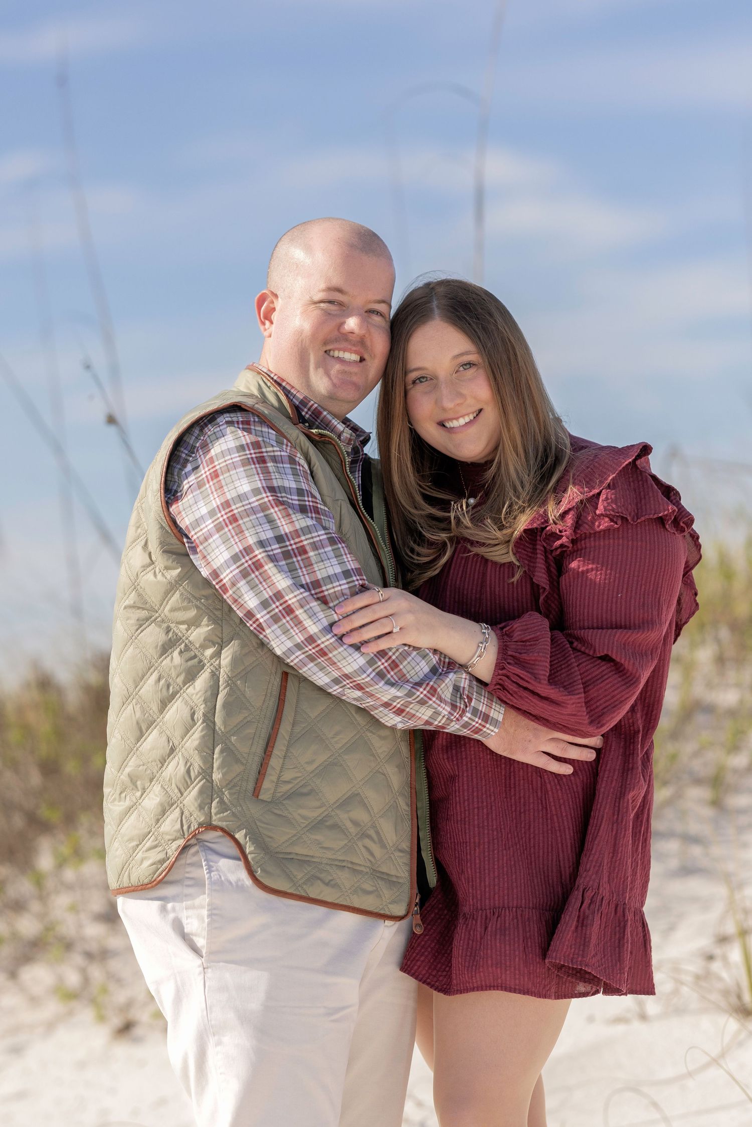 Happy couple embracing on a sandy beach, man in quilted vest and woman in burgundy dress, with sea oats and blue sky.