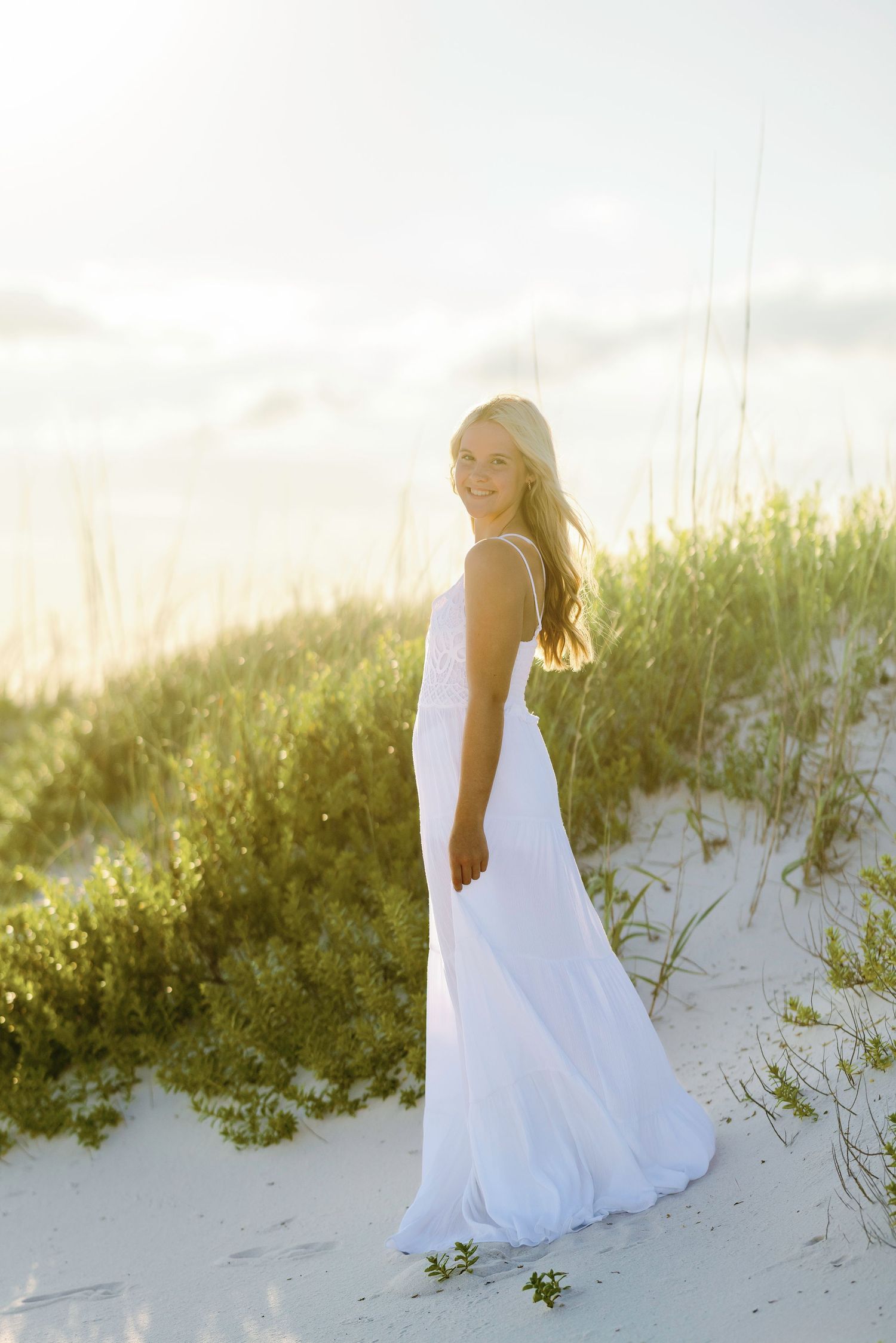 Young woman in white maxi dress standing on sandy beach dunes at golden hour sunset.