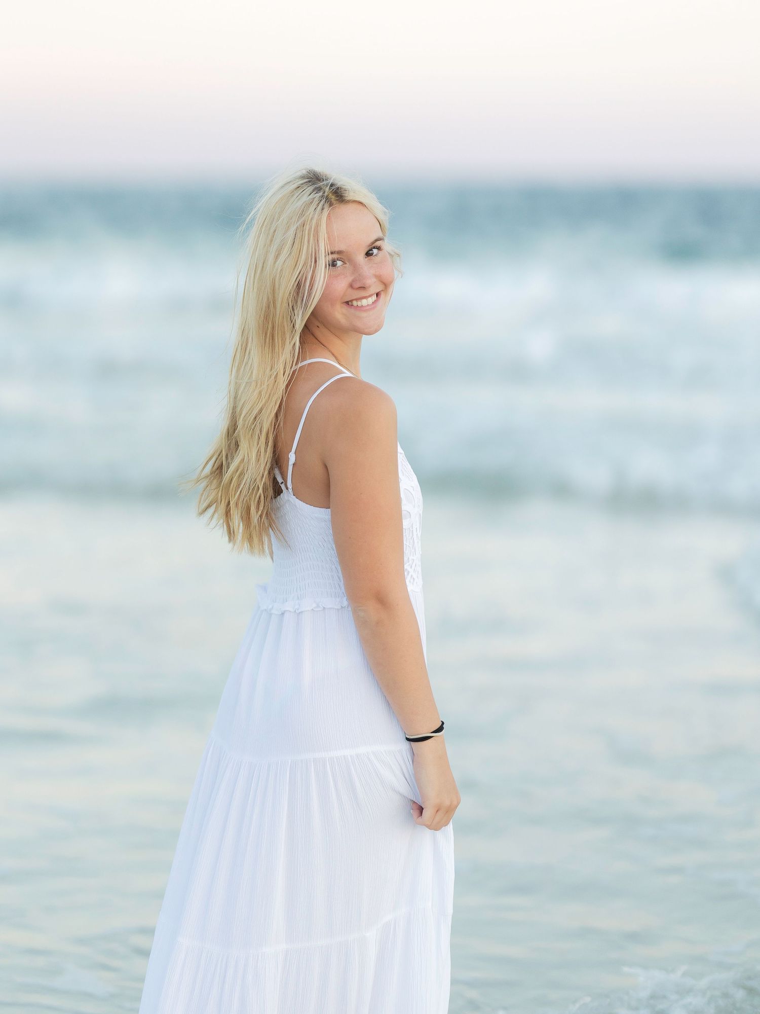 Smiling blonde woman in white sundress posing on beach with ocean waves behind her.