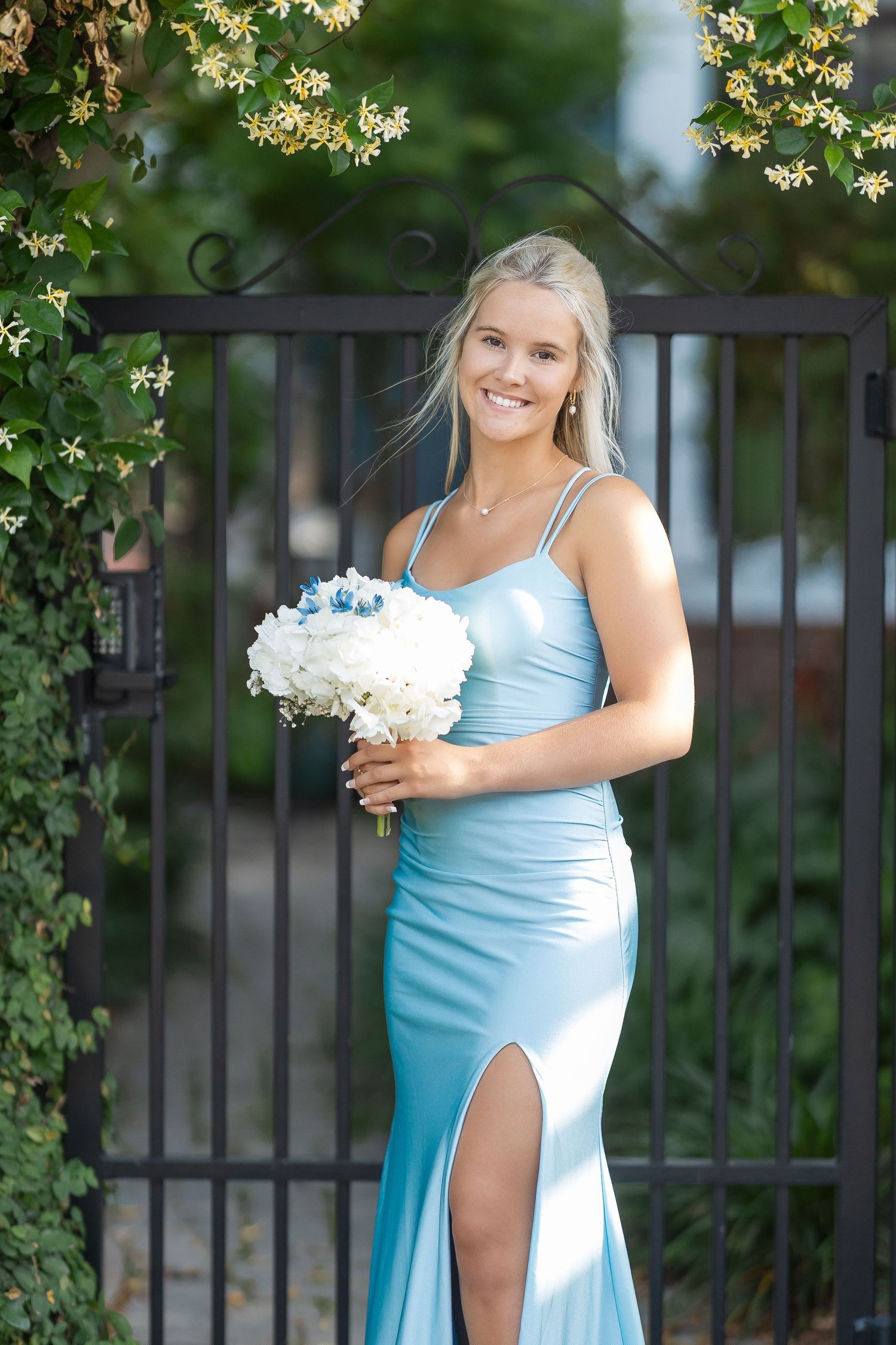 Young blonde woman in a light blue satin gown holding white bouquet, smiling by an iron gate with lush greenery.