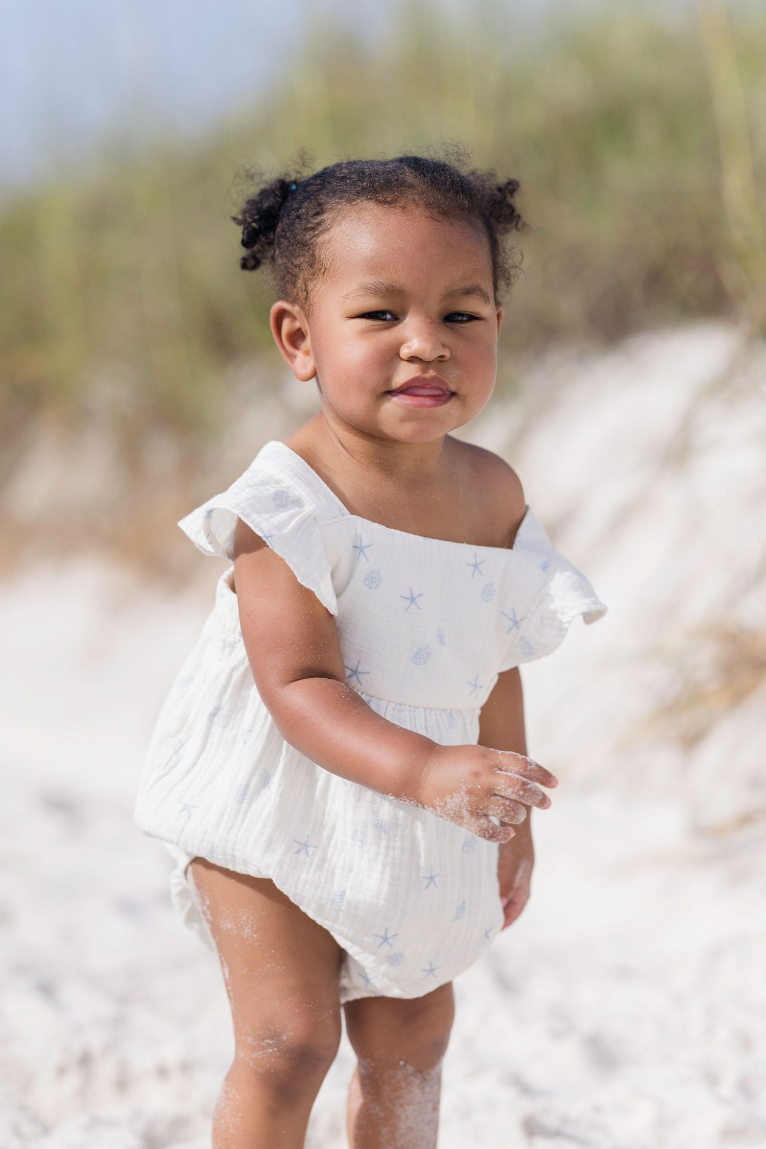 Toddler girl in white floral romper playing on sandy beach, smiling outdoors on a sunny day.