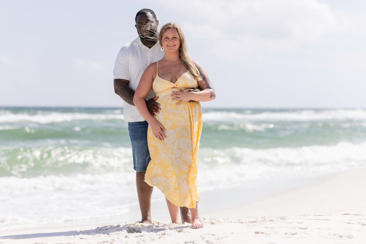 Pregnant interracial couple standing on a beach, woman in yellow dress cradling baby bump, man embracing her from behind.