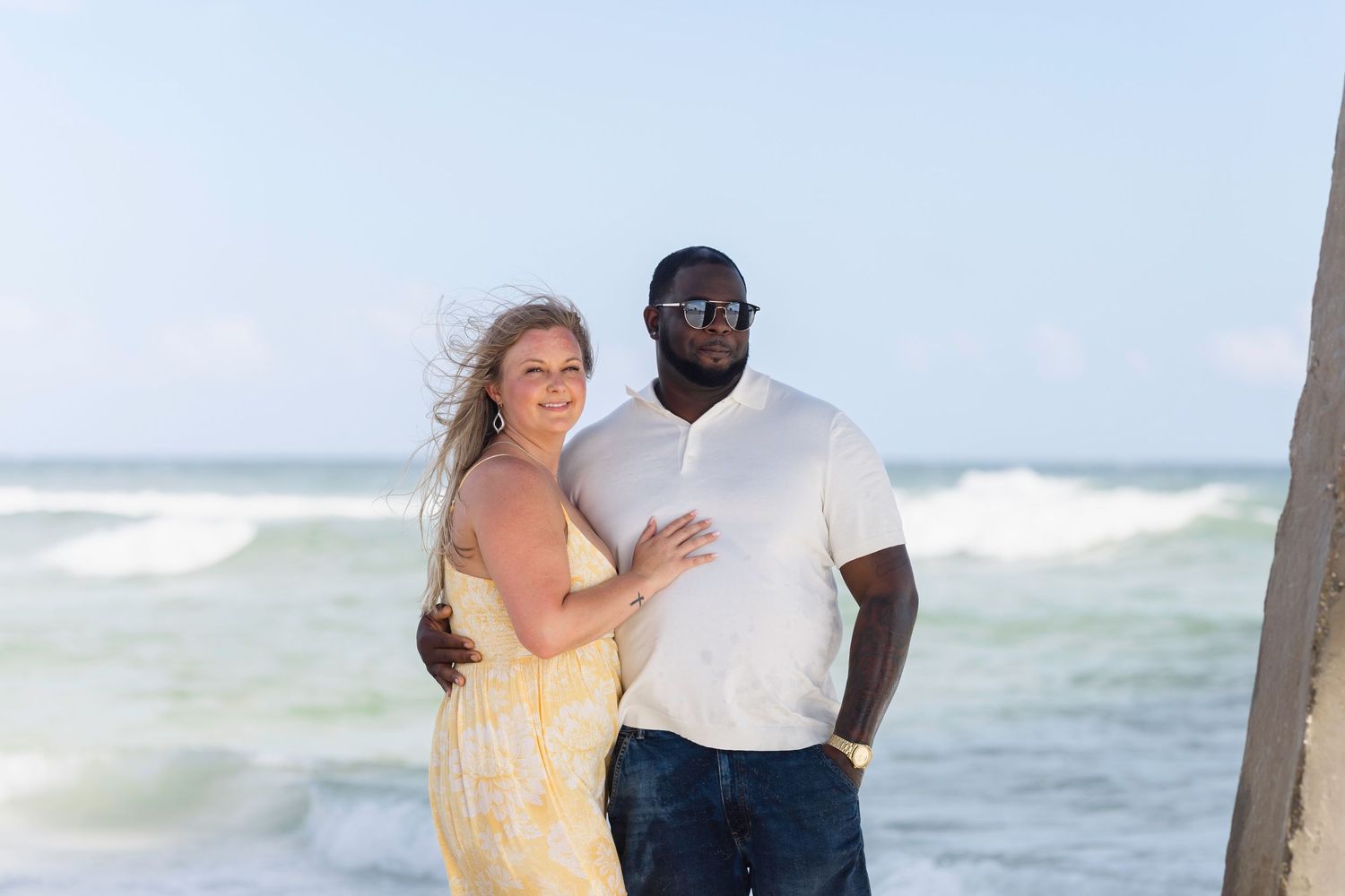 Couple posing on a beach, woman in yellow dress and man in white polo shirt, with ocean waves in the background.