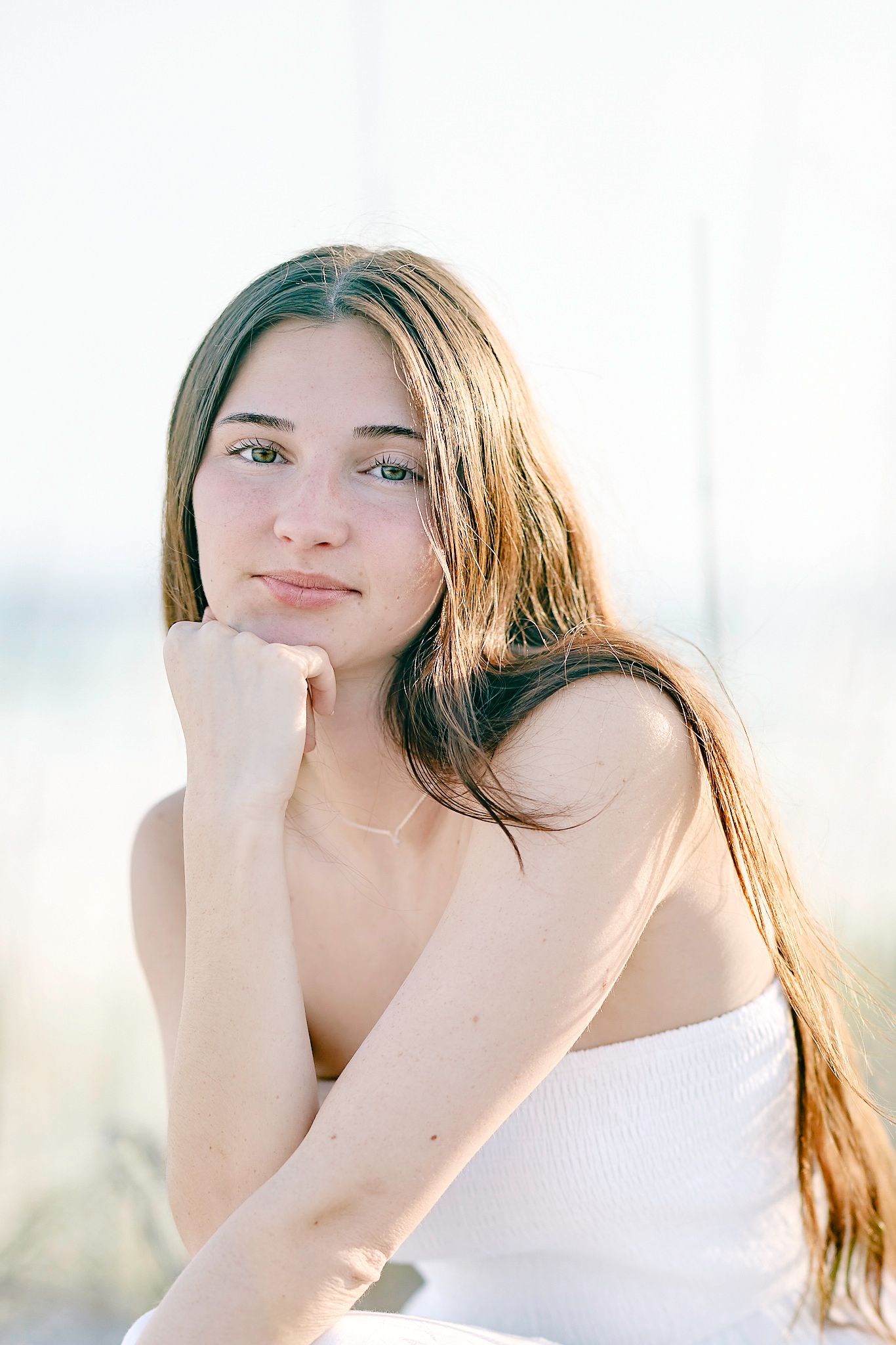 Young woman with long brown hair in white top resting chin on hand in bright natural light.