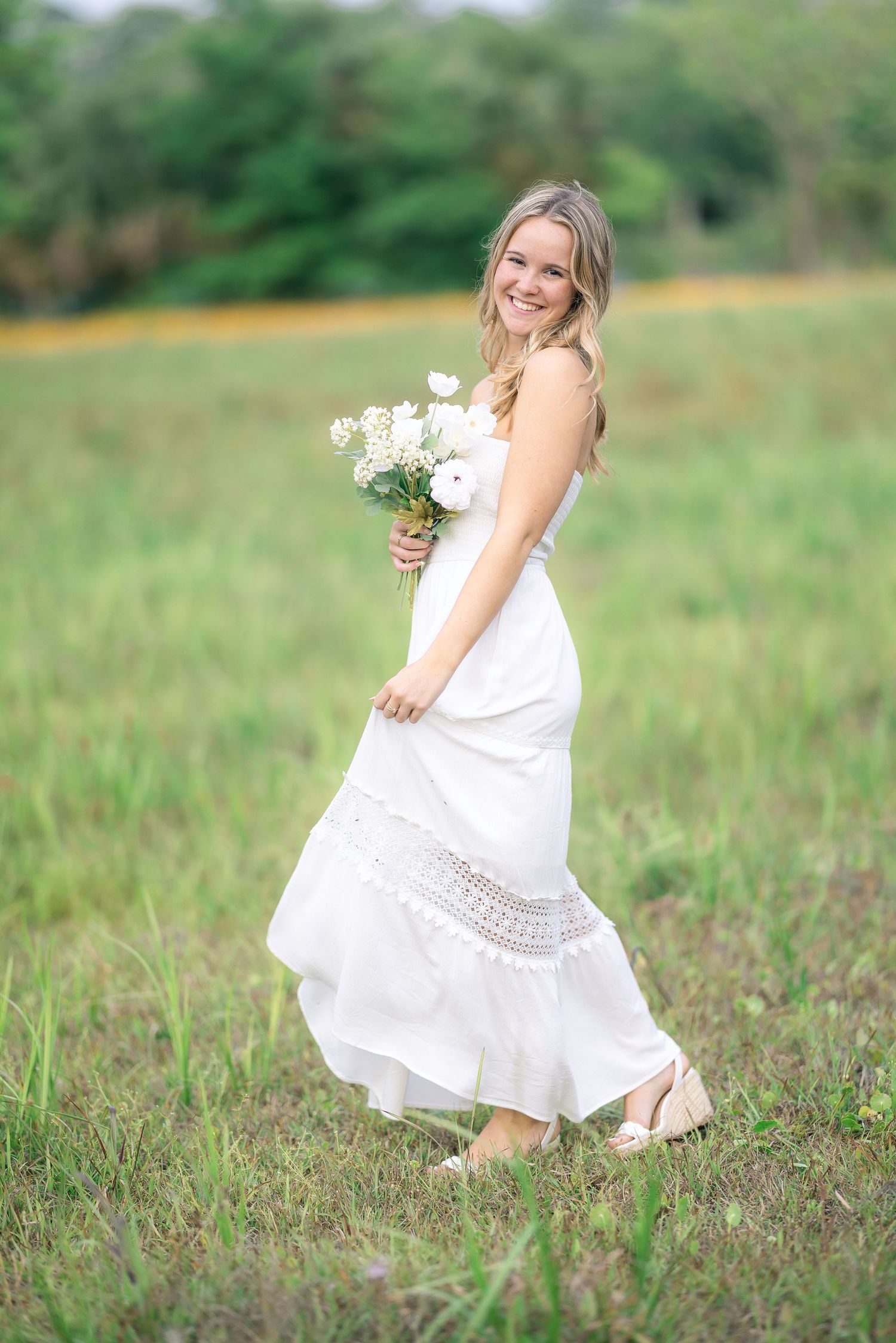 Happy blonde woman in white boho maxi dress holding white flower bouquet while walking through lush green field.