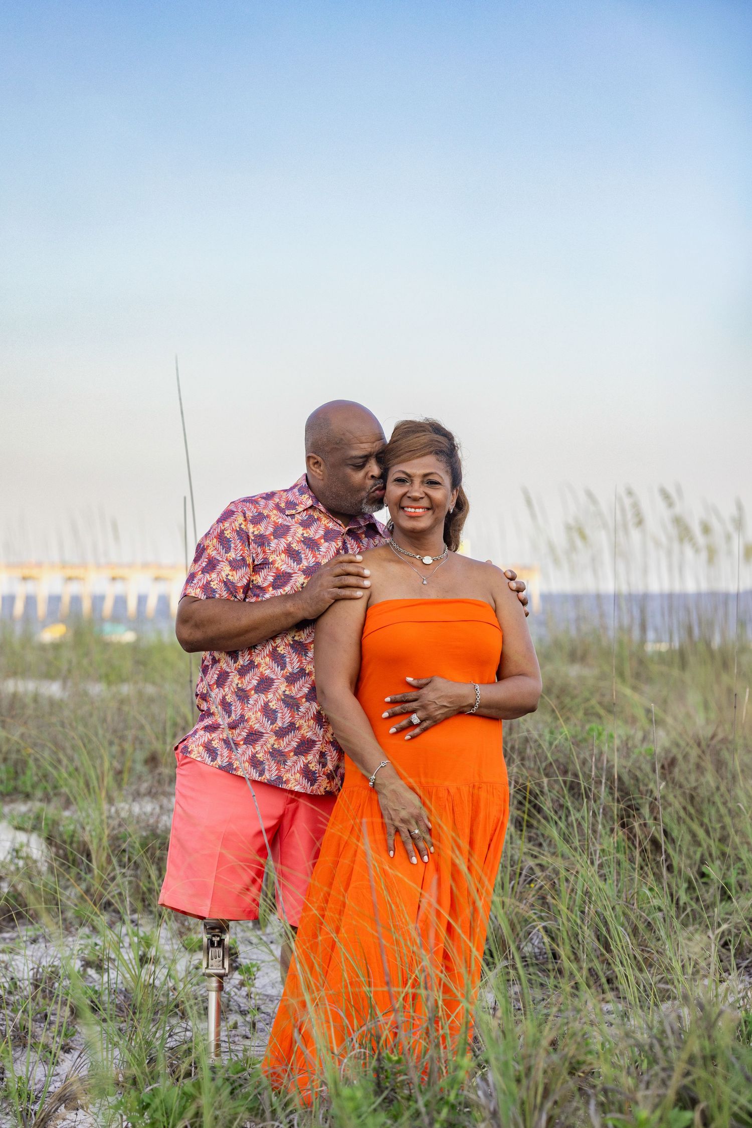Couple posing on beach dunes, man in floral shirt kissing woman in orange dress at sunset.