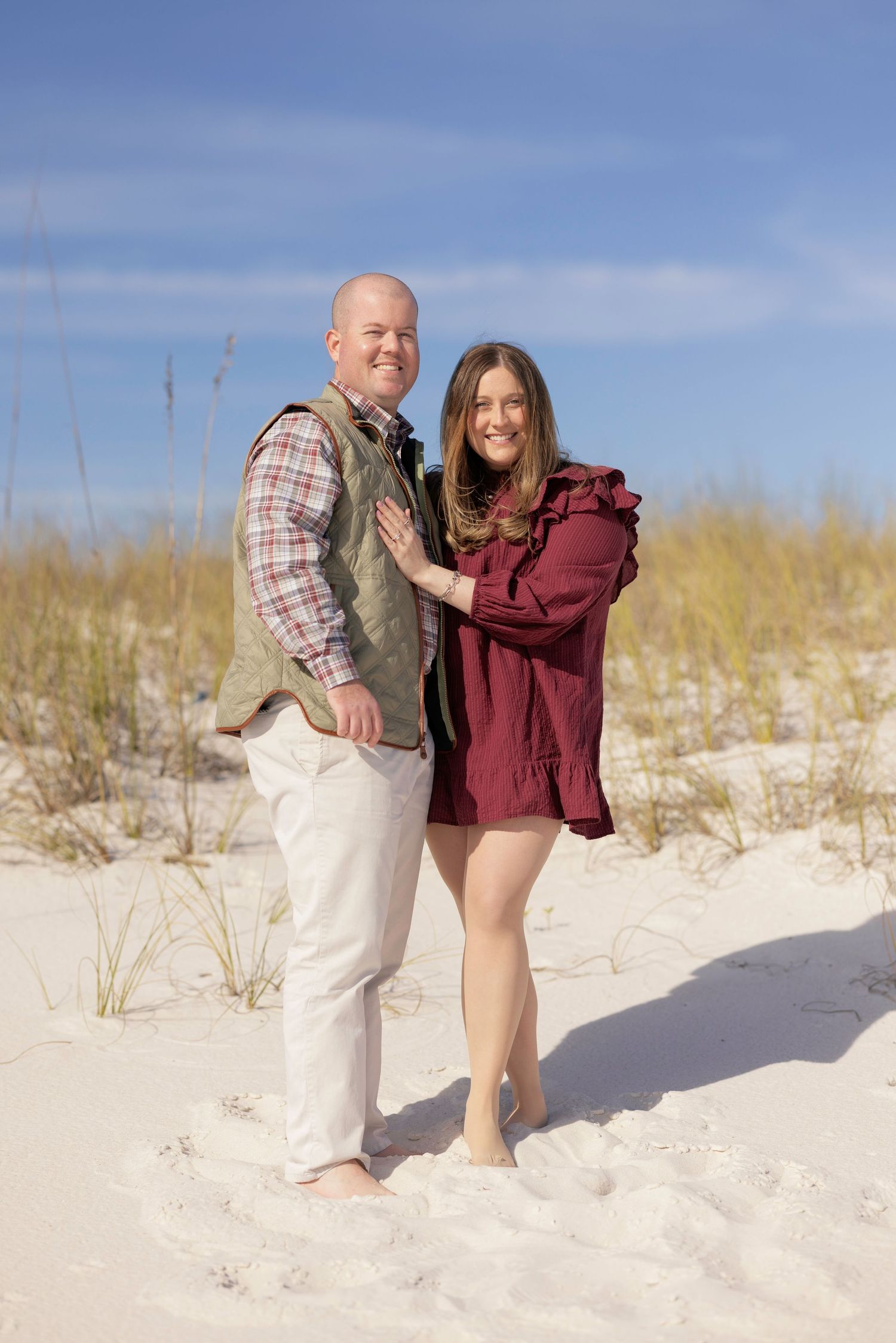 Smiling couple posing together on white sand dunes, man in green quilted vest and woman in burgundy ruffled dress.