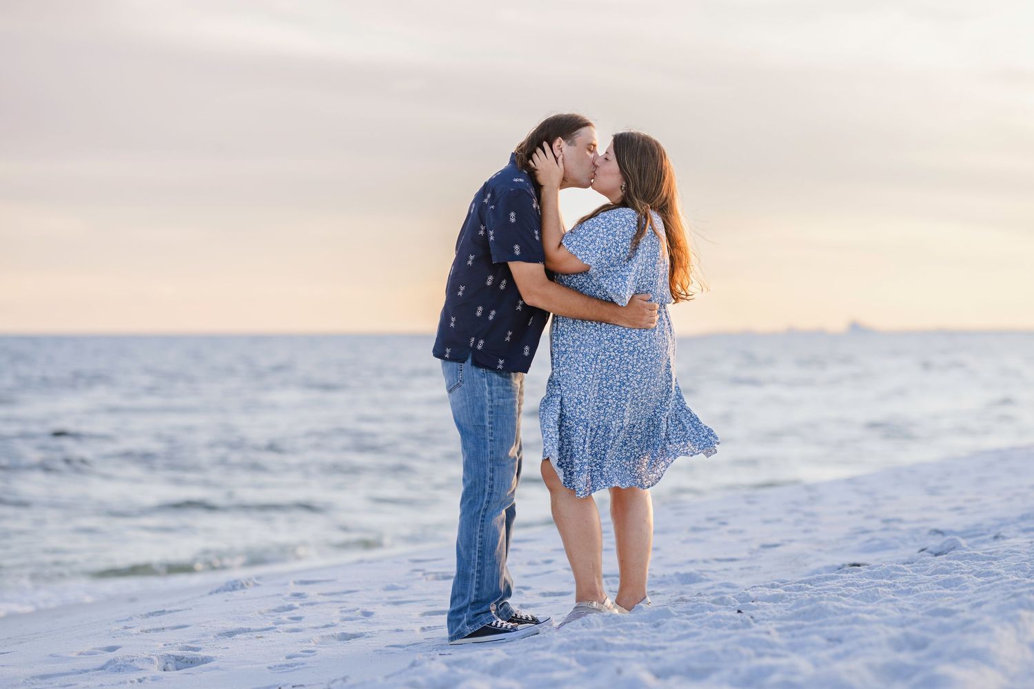 Couple sharing a romantic kiss on a sandy beach at sunset, woman in blue floral dress, man in dark shirt and jeans.
