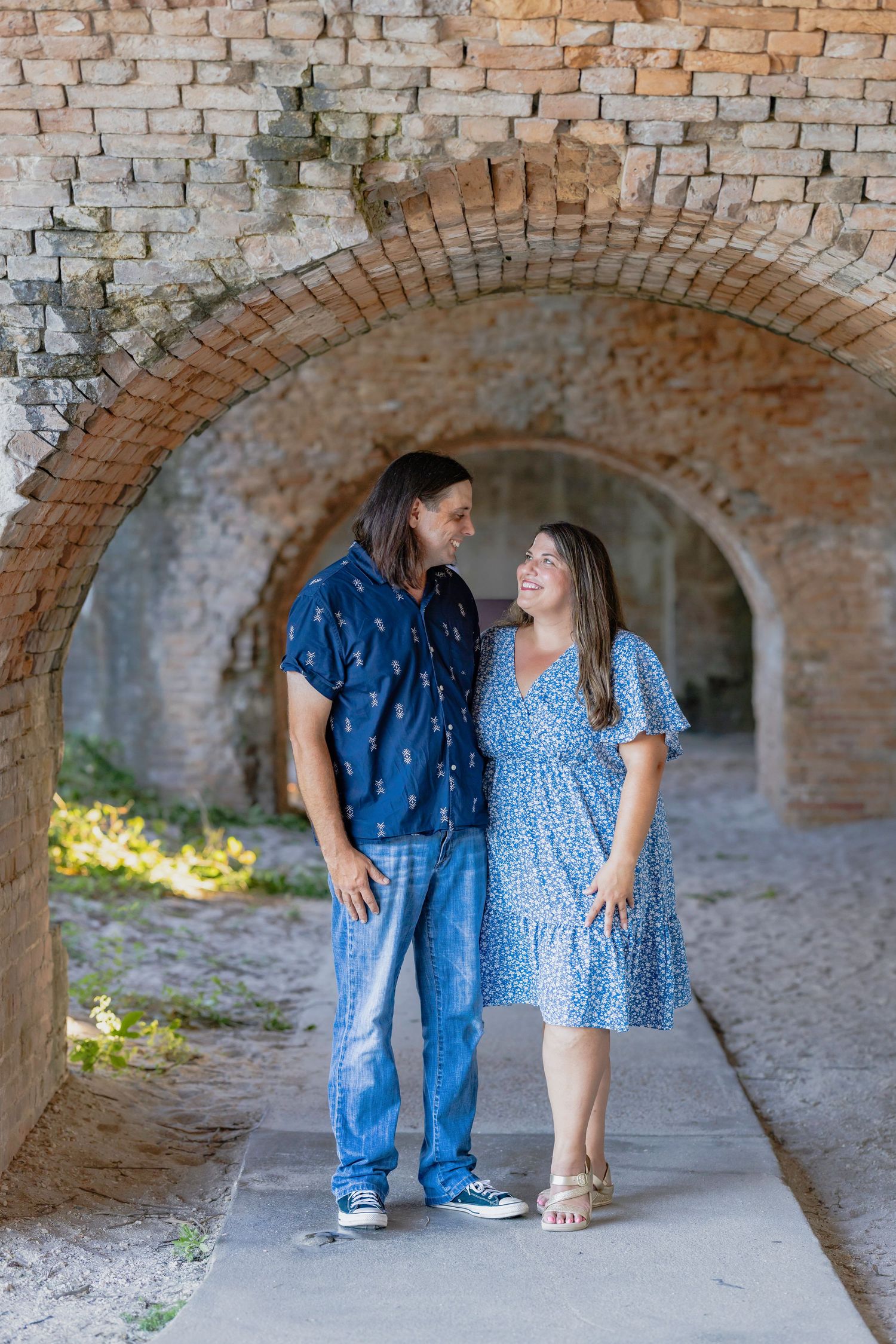 Couple smiling at each other under brick arches, woman in blue floral dress, man in blue shirt and jeans.