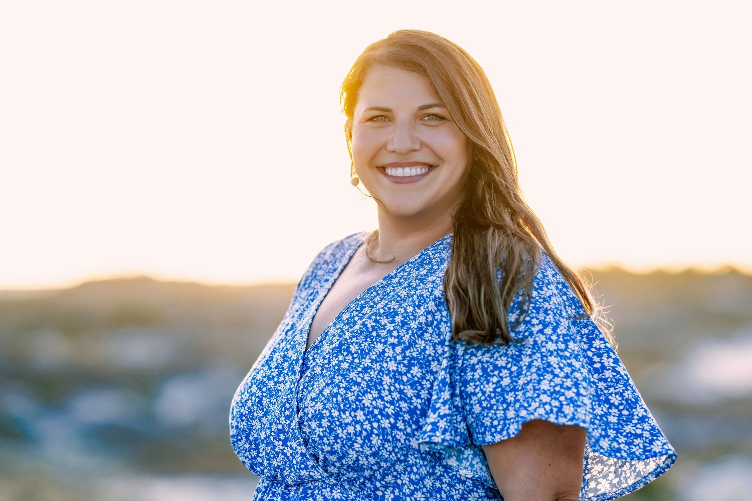 Smiling woman in blue floral dress at golden hour sunset, long auburn hair, outdoor portrait photography.