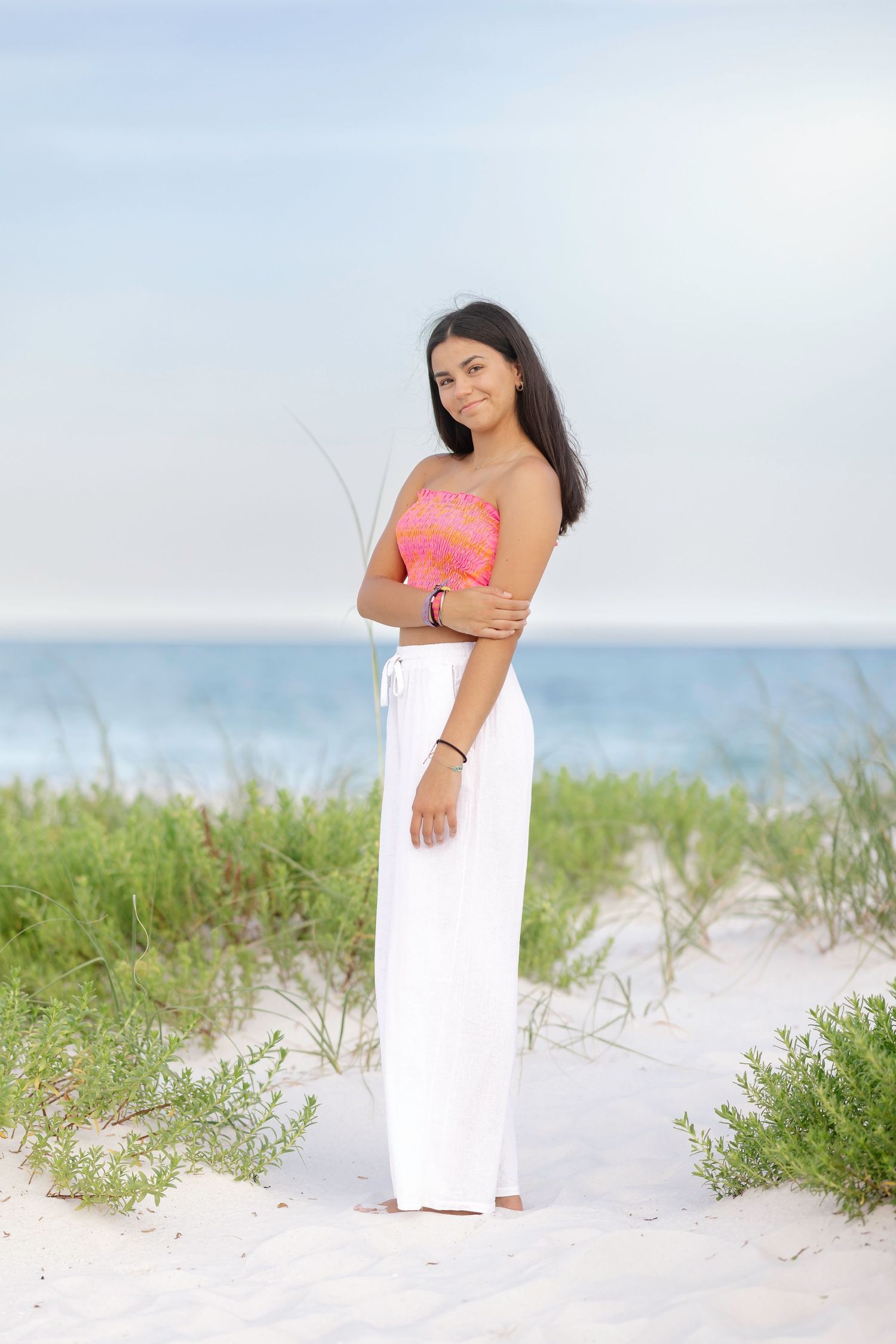 Woman in flowing white skirt and coral top stands gracefully on sandy beach.