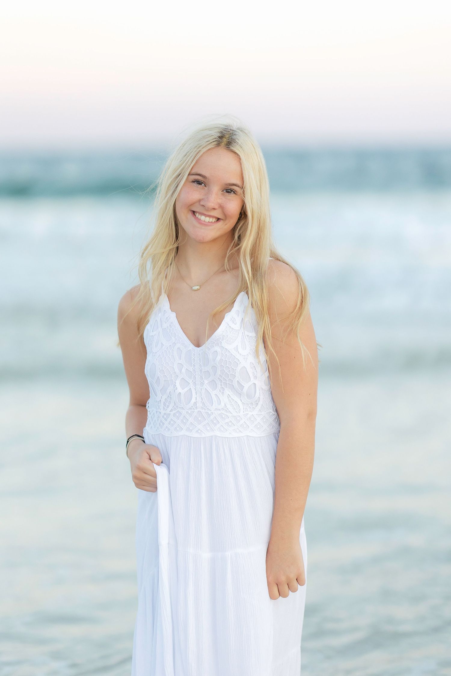 Woman in a white lace maxi dress smiles while posing on the beach at sunset.