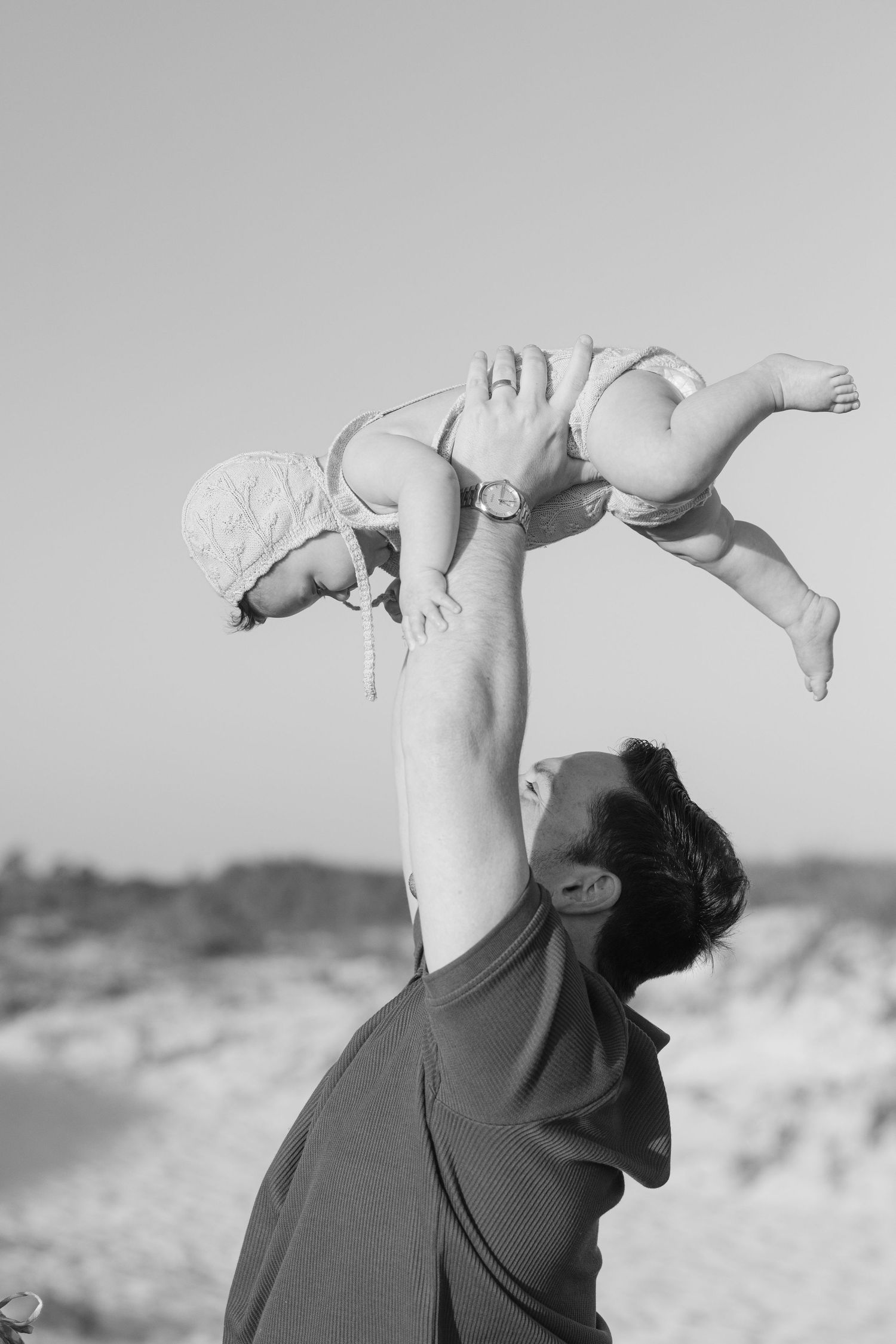 Black and white photo of a father lifting his baby overhead on a beach, sharing a joyful moment.