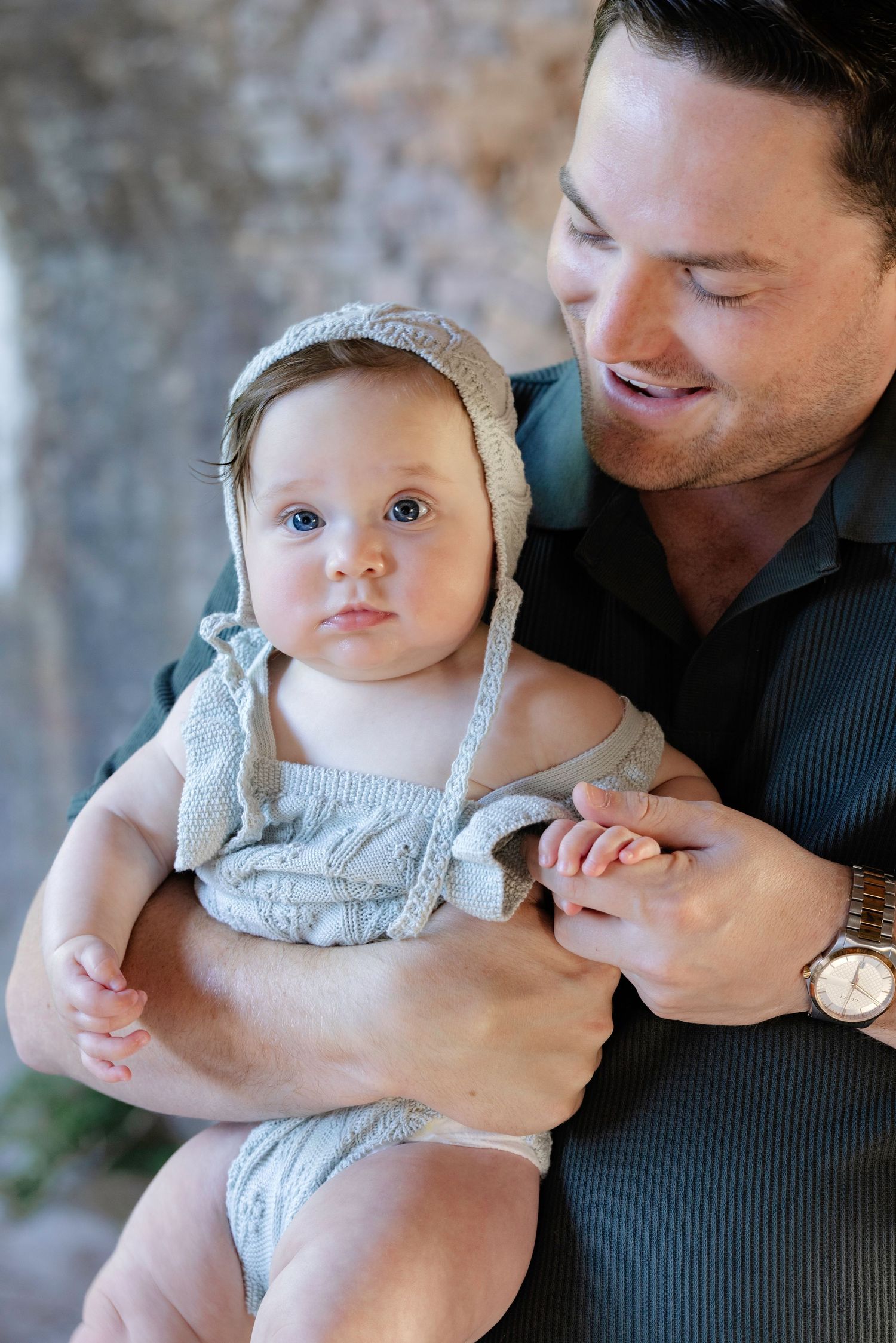 Smiling man holding an adorable baby in a knit bonnet and romper, sharing a tender moment together.