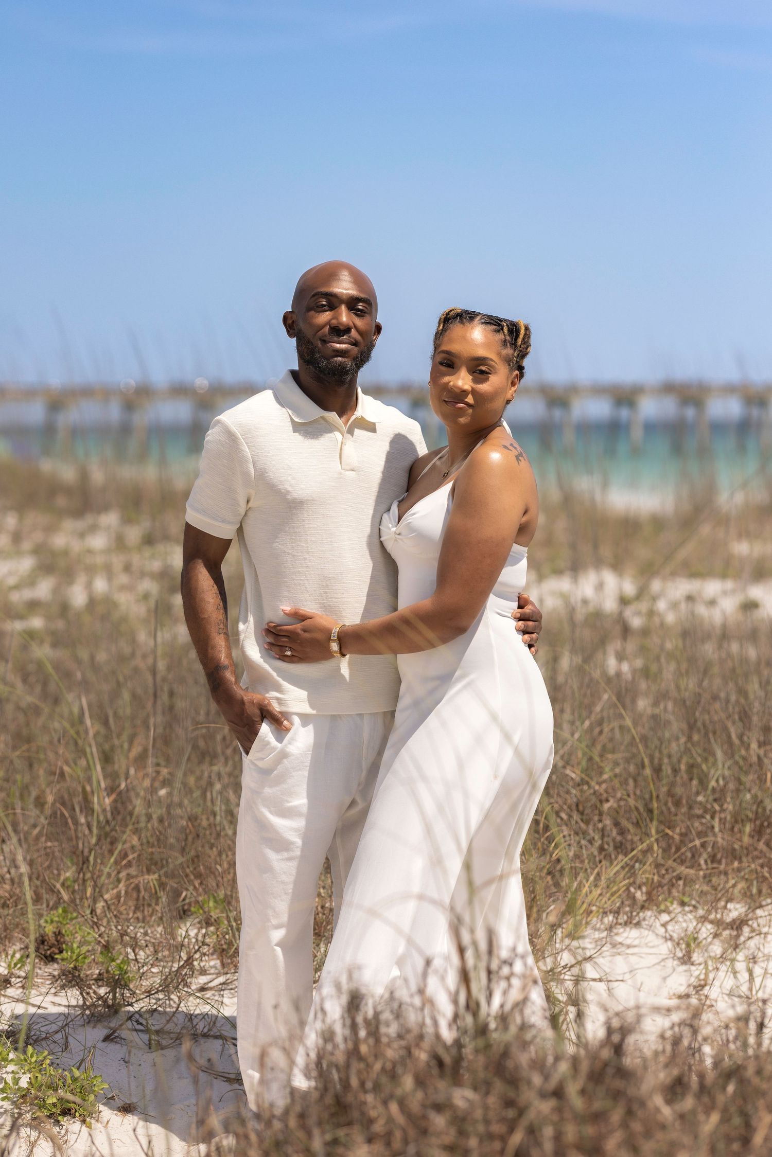 A couple in matching white outfits poses on a sandy beach with ocean and pier in the background on a sunny day.
