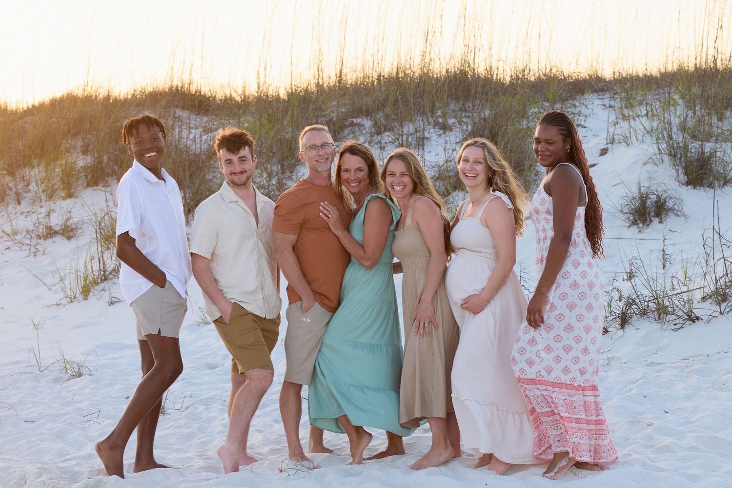 Family of seven smiling together on white sand beach at sunset with sand dunes behind them.