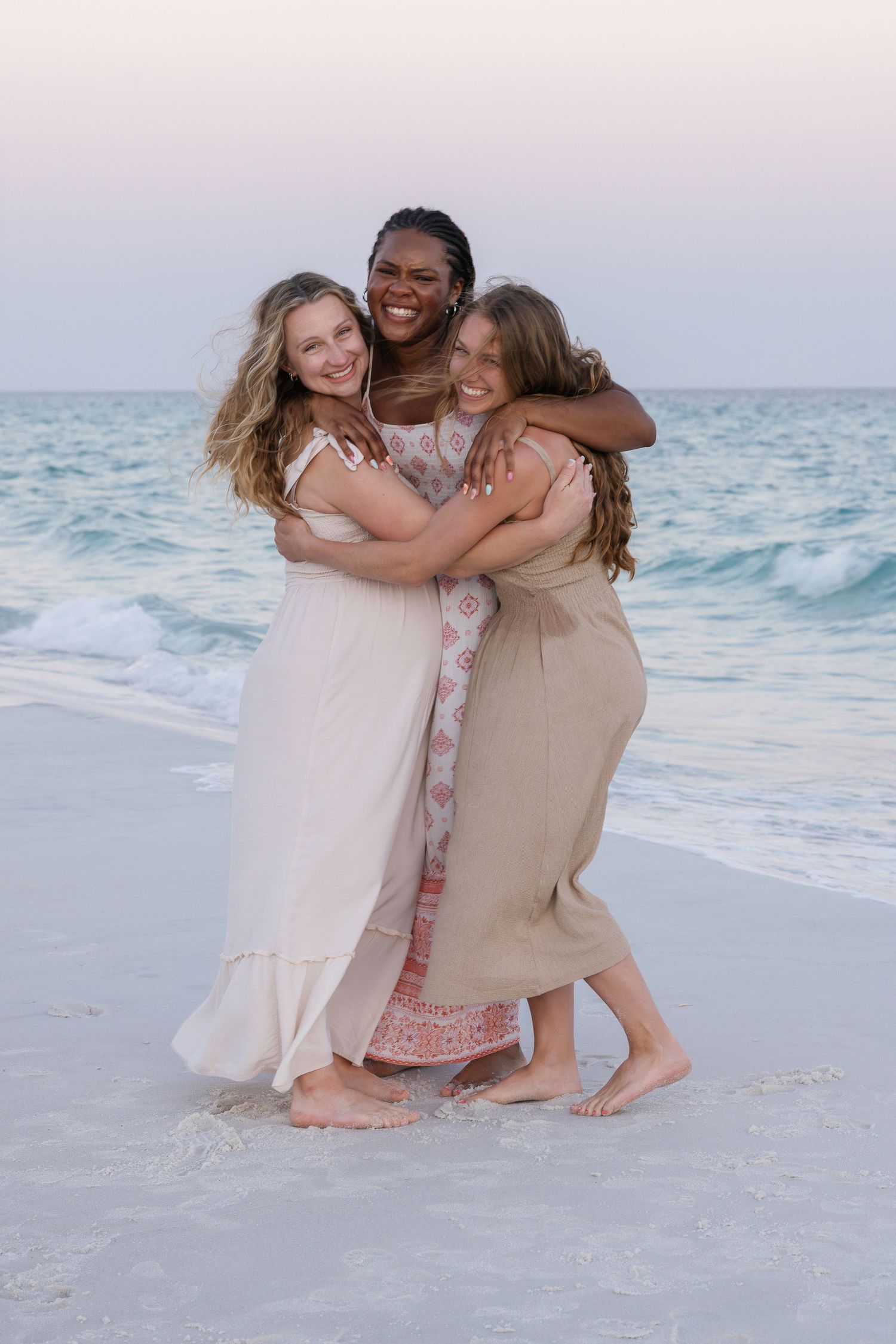 Three women in summer dresses share a joyful group hug on a beach at sunset.