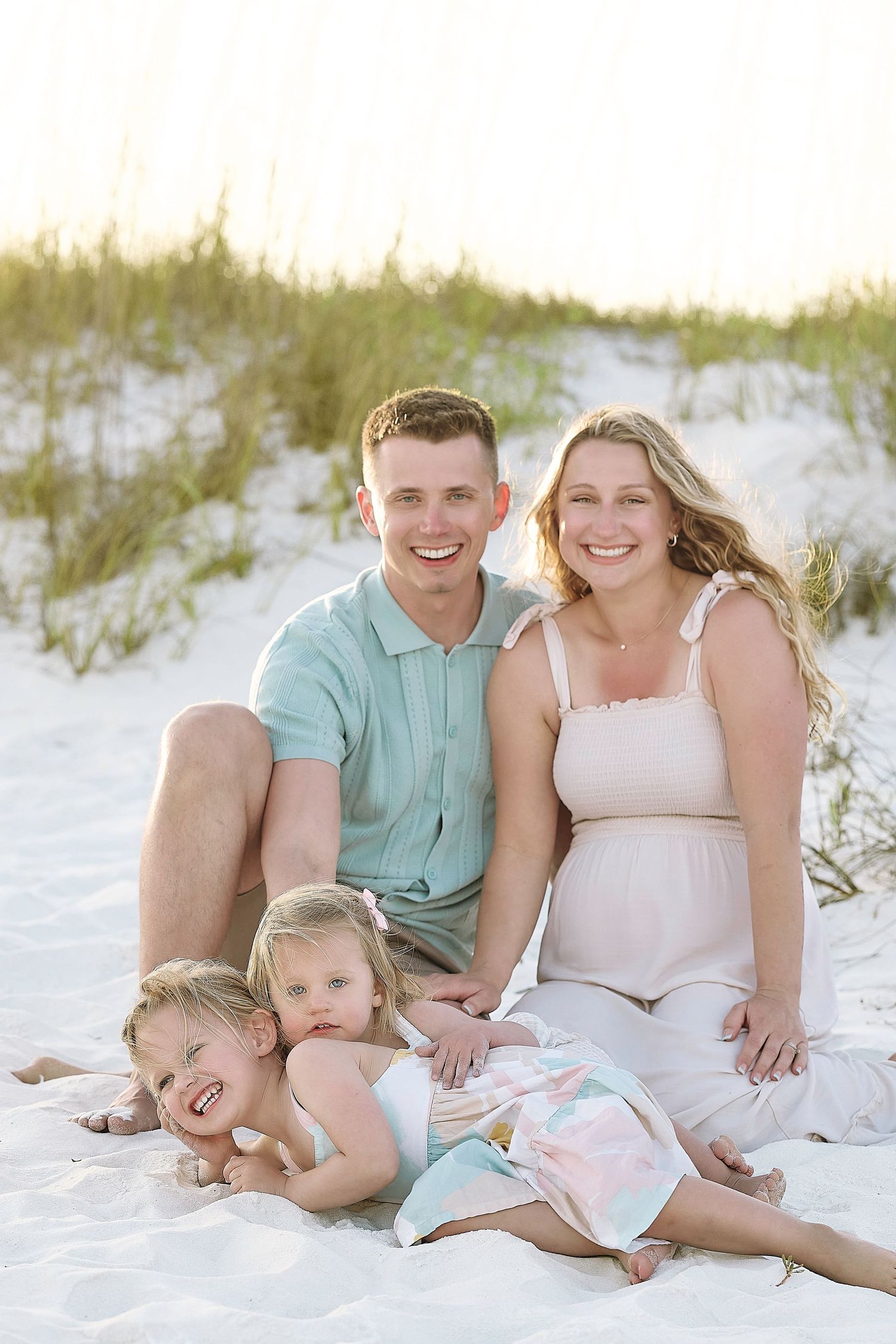 Pregnant woman and man smile with two young children on white sand beach dunes during golden hour family photo session.
