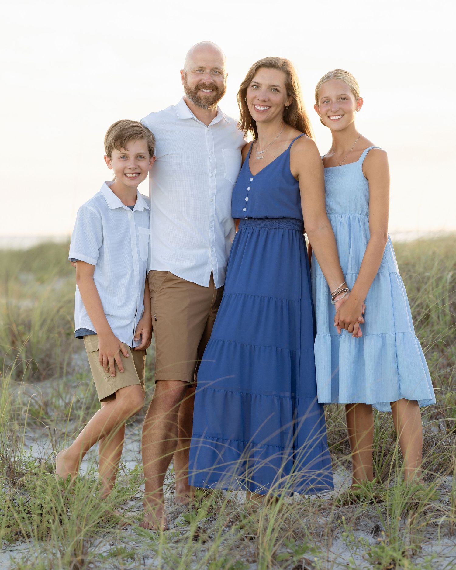Family of four posing together on beach dunes at sunset, wearing coordinated blue and khaki outfits.