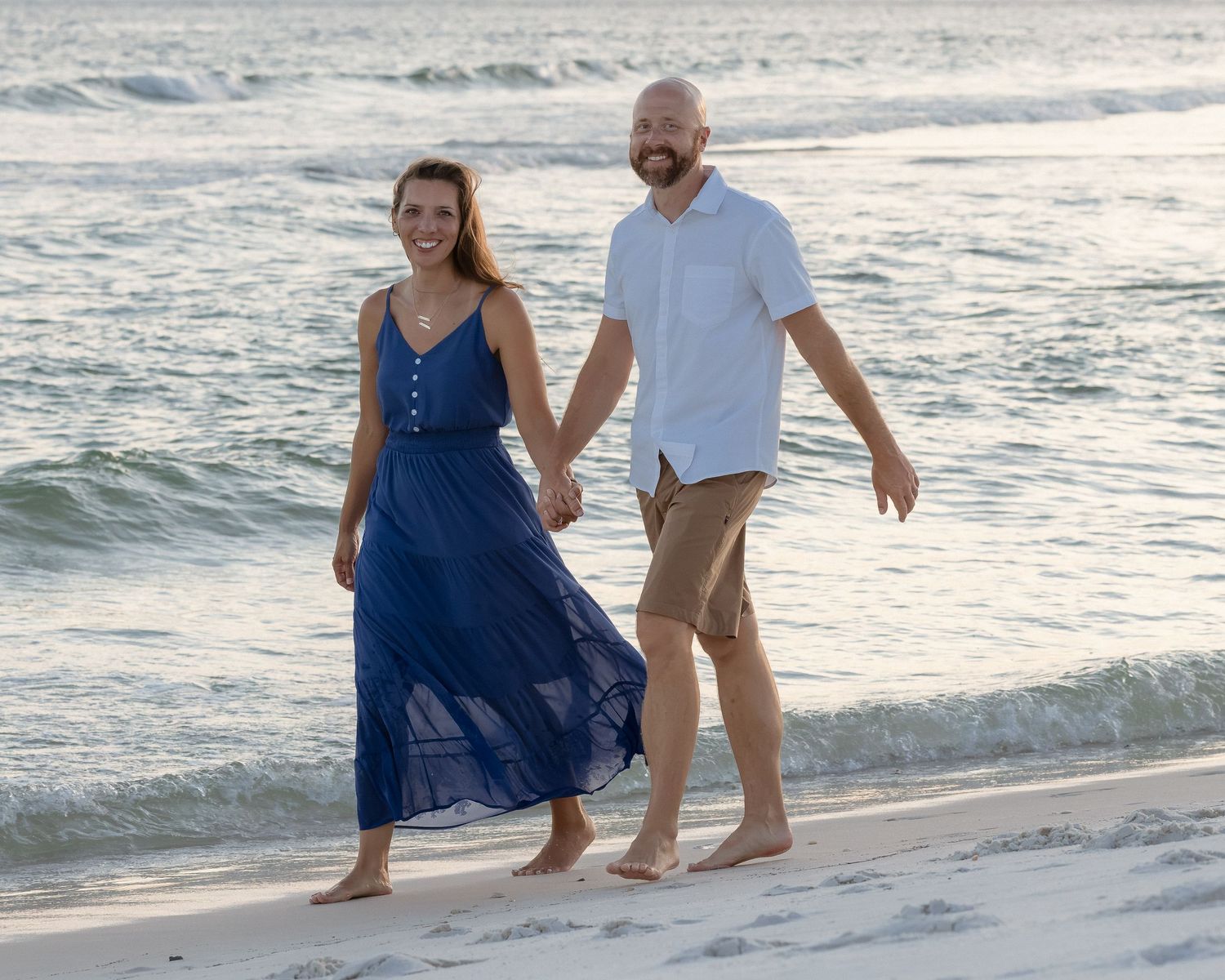 Couple holding hands walking barefoot on a sandy beach, woman in blue maxi dress, man in white shirt and khaki shorts.