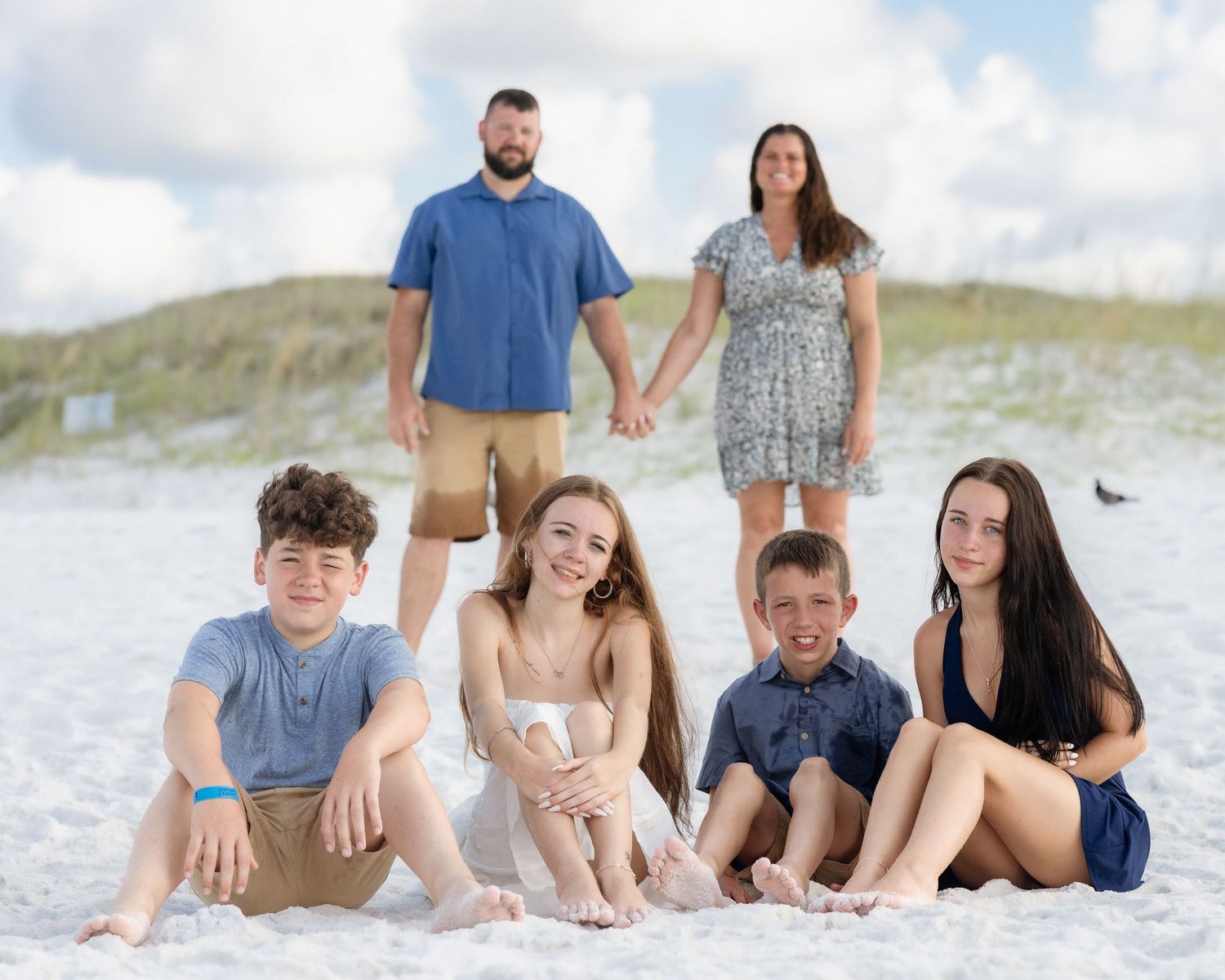 Family of six posing on white sand beach, parents holding hands behind four teens sitting in sand.