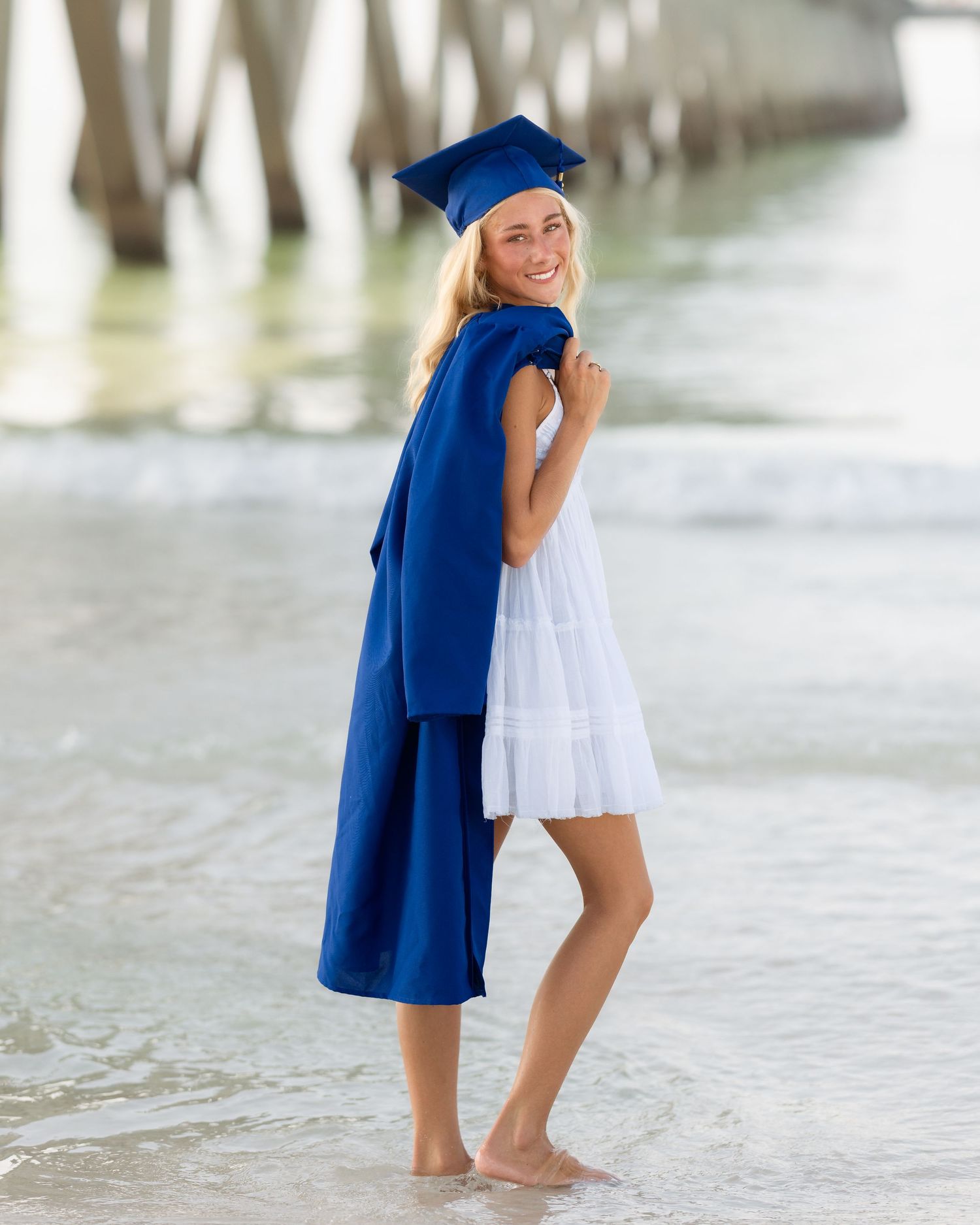 Smiling graduate in blue cap and gown over white dress standing barefoot on beach shoreline.