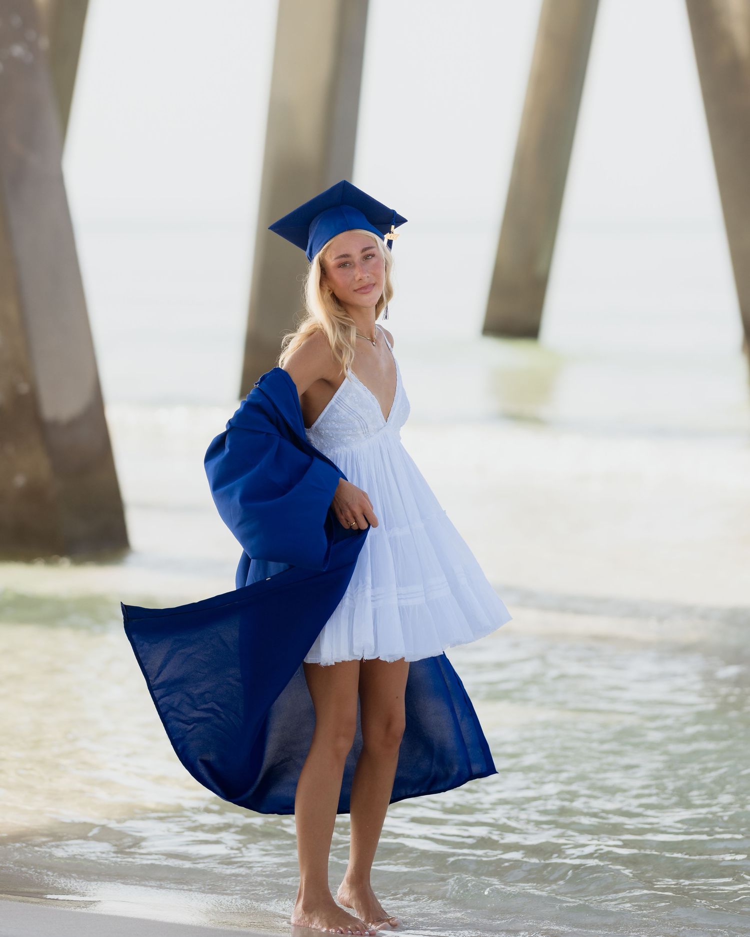 Smiling graduate in blue cap and gown over white dress standing barefoot on beach near pier.