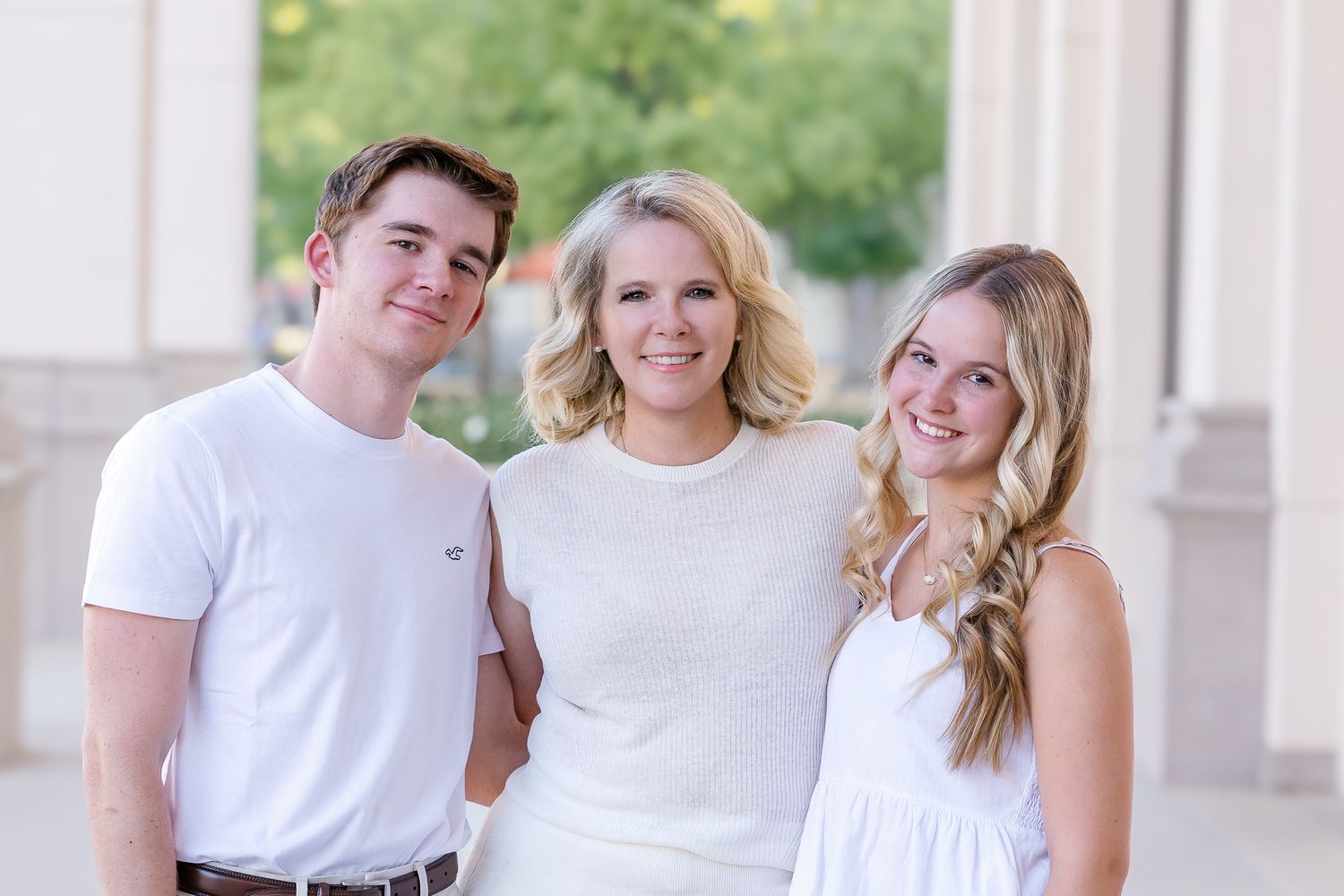 Three smiling people in white outfits pose together outdoors near elegant columns in a bright, cheerful photo.