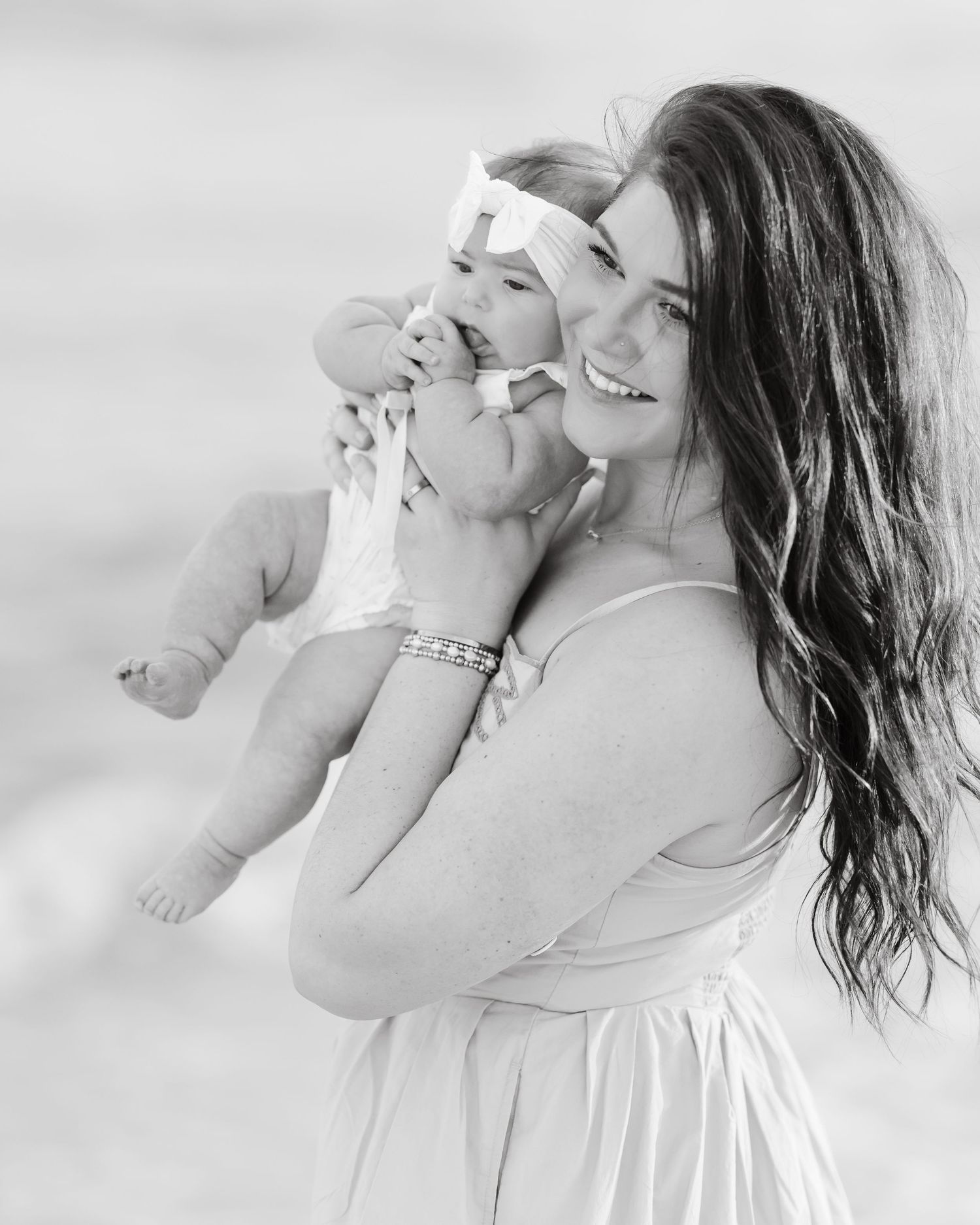 Black and white photo of a smiling young mother holding her laughing baby girl wearing a bow headband outdoors.