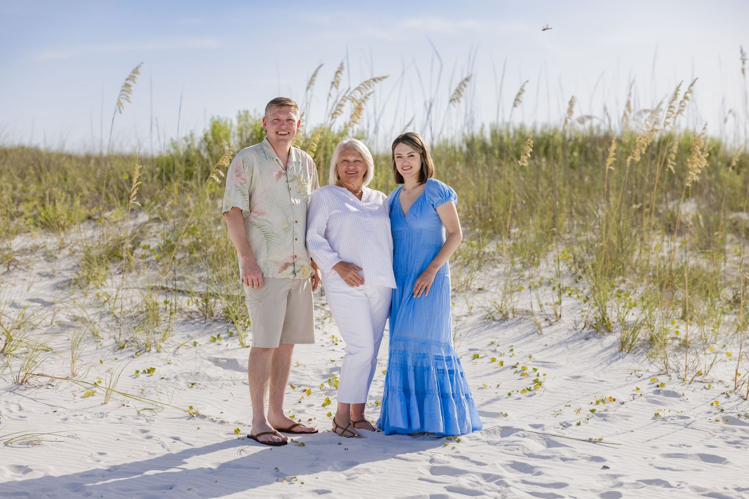 Family of three posing together on a sandy beach with sea oats and blue sky in the background.