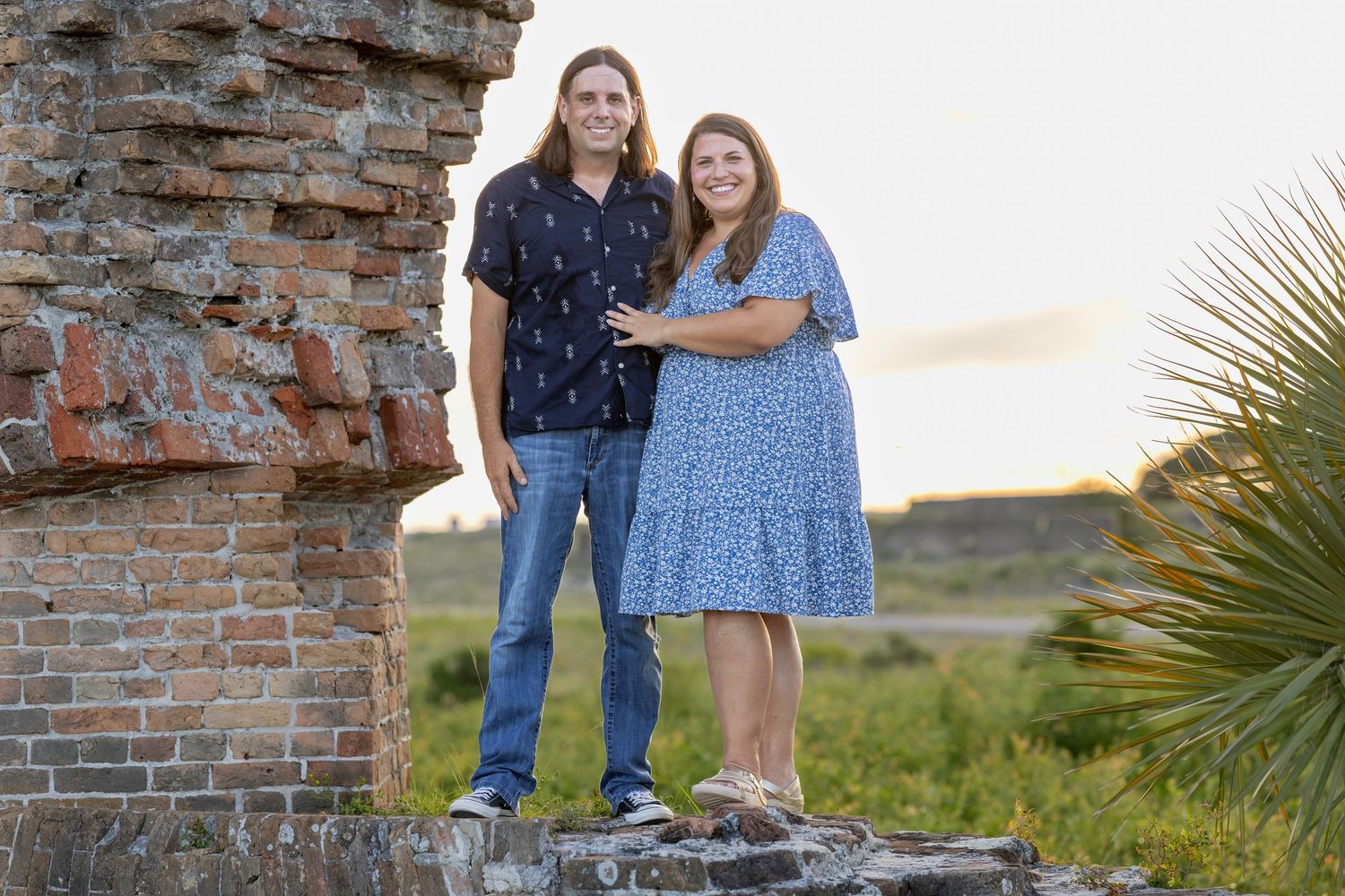Happy couple standing on ruins at sunset, woman in blue dress and man in dark shirt with jeans.