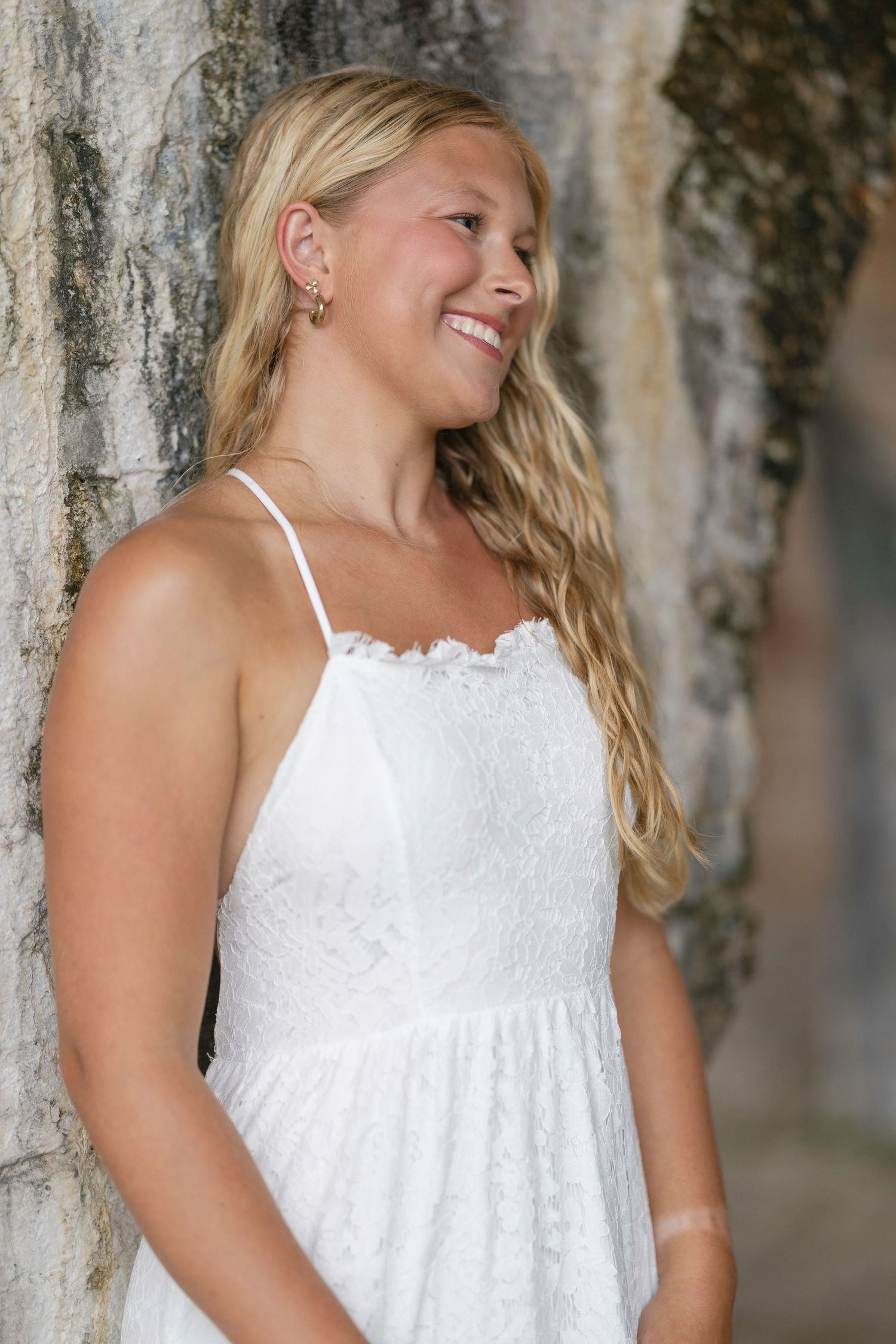 Smiling blonde woman in white lace sundress leaning against stone wall outdoors.
