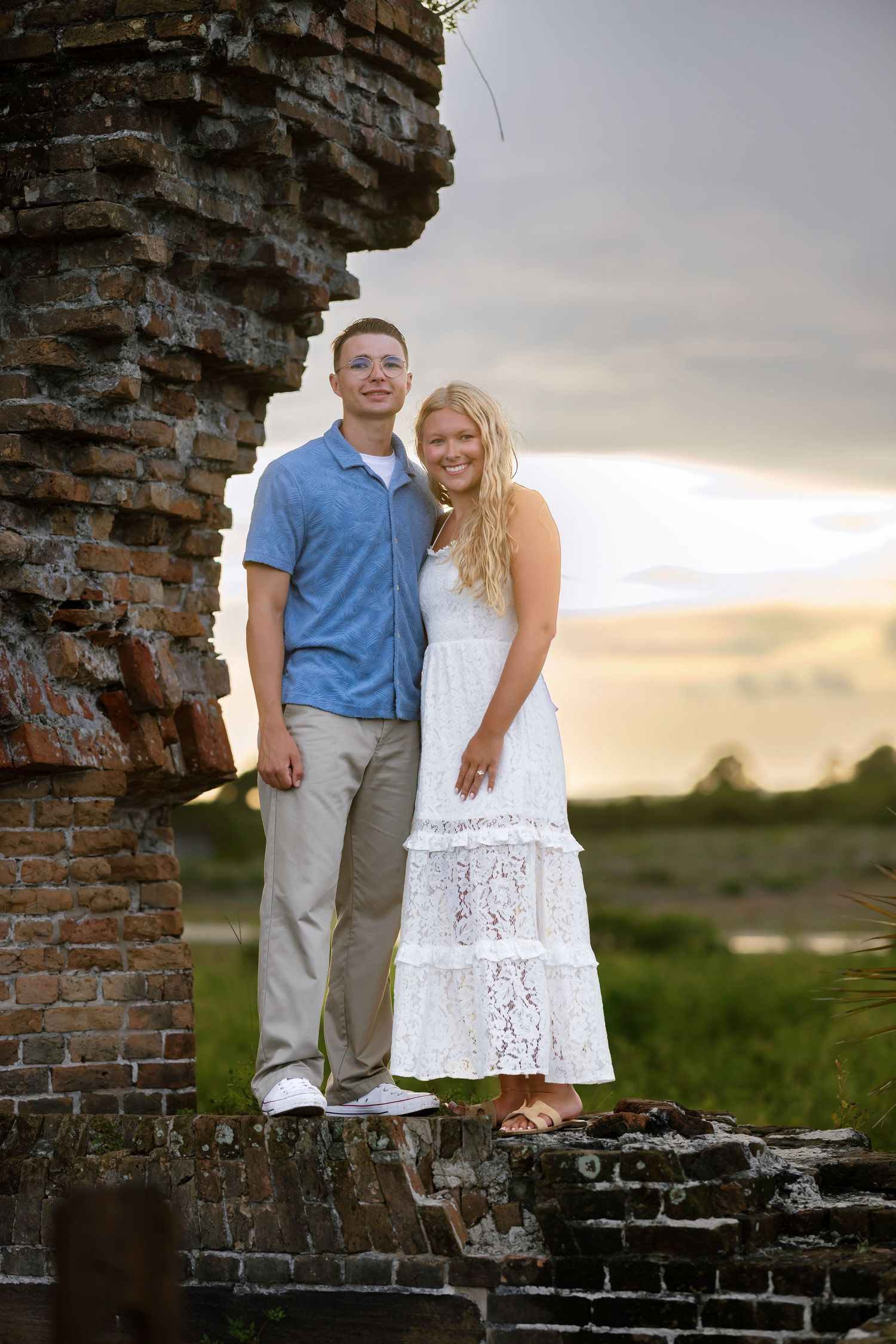 A smiling couple poses together on historic brick ruins at sunset, woman in white lace dress and man in blue shirt.
