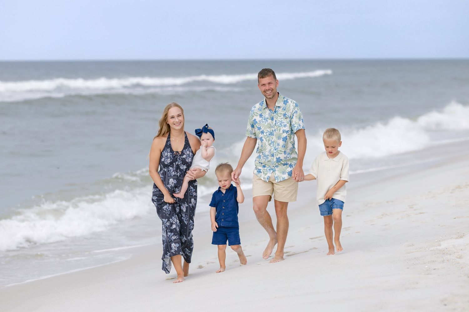 Happy family of five walking along a sunny beach, waves in background, wearing coordinated outfits.