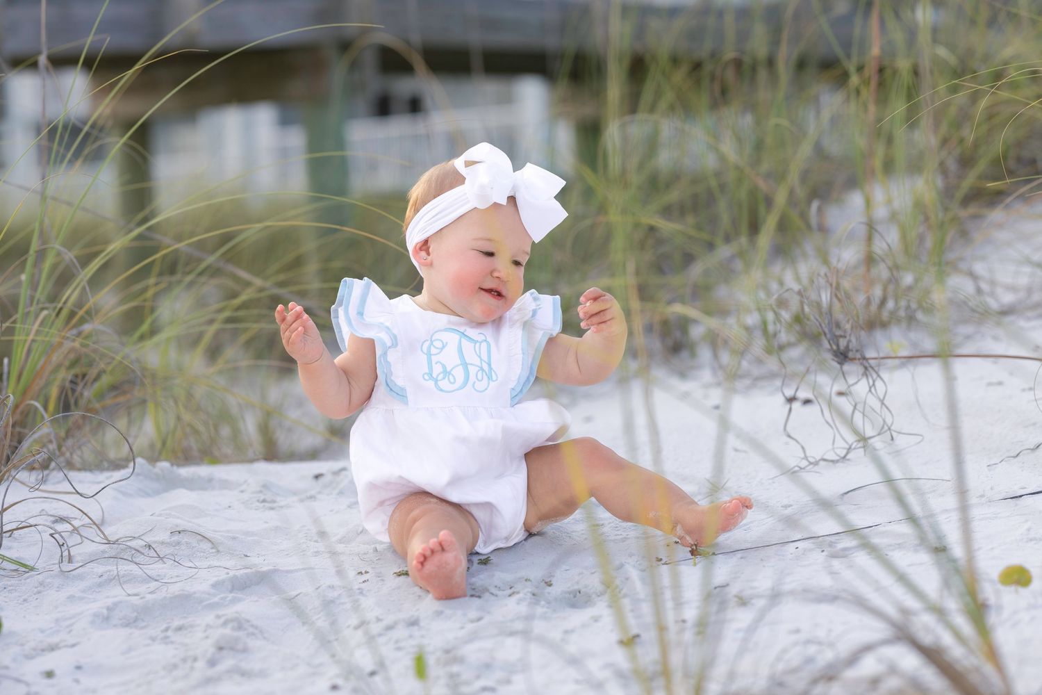 Baby girl in white monogrammed romper with bow headband sitting on white sand beach surrounded by sea grass.