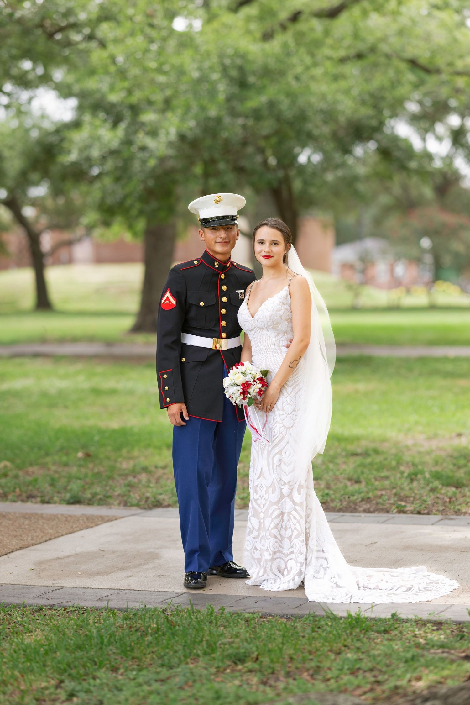 Marine in dress uniform stands with bride in white lace gown holding bouquet, posing outdoors on park path.