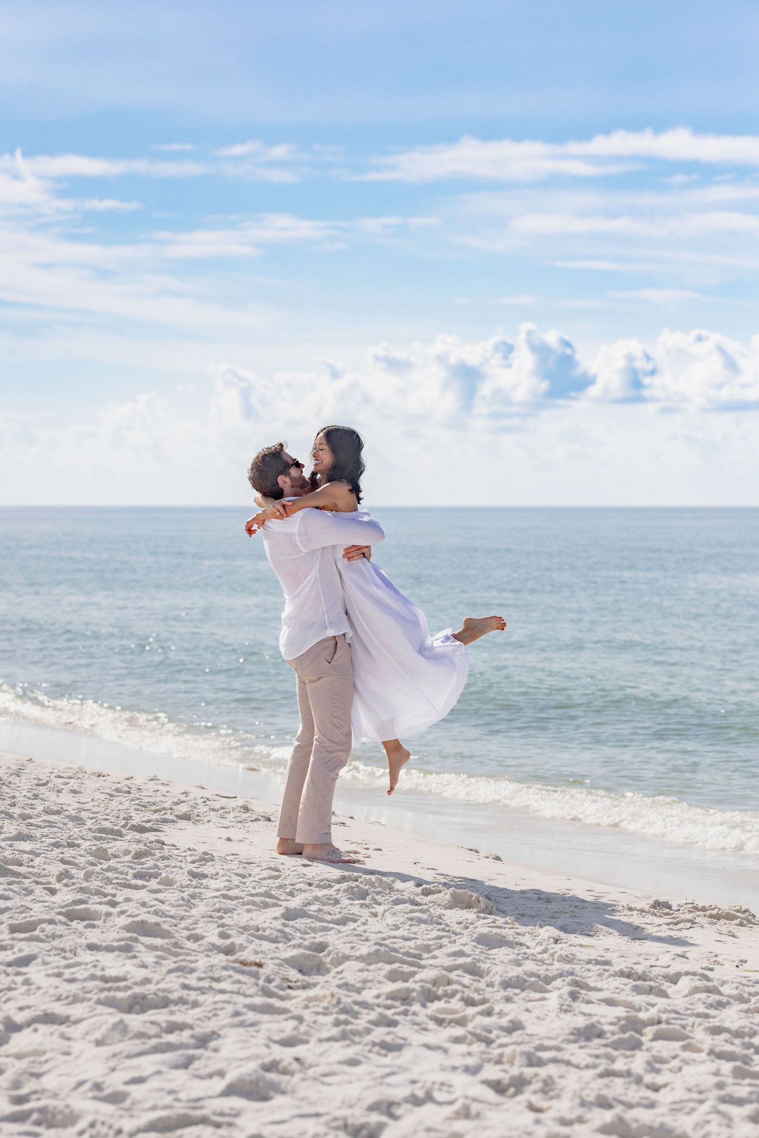 Romantic couple embracing on a white sandy beach, man lifting woman in white dress, ocean waves and blue sky behind them.
