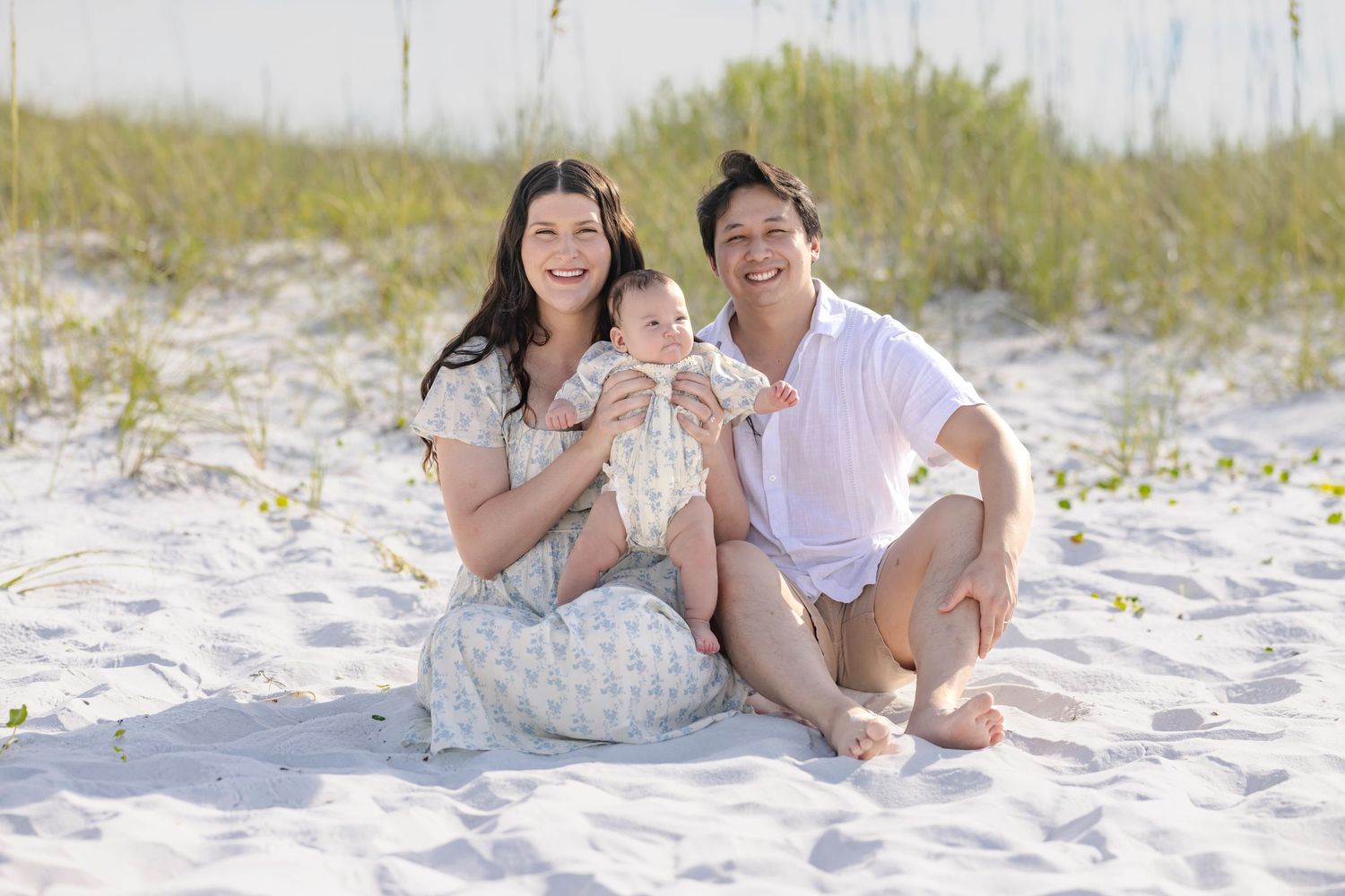 Happy family with newborn baby sitting on white sand beach, smiling couple enjoying sunny day outdoors.
