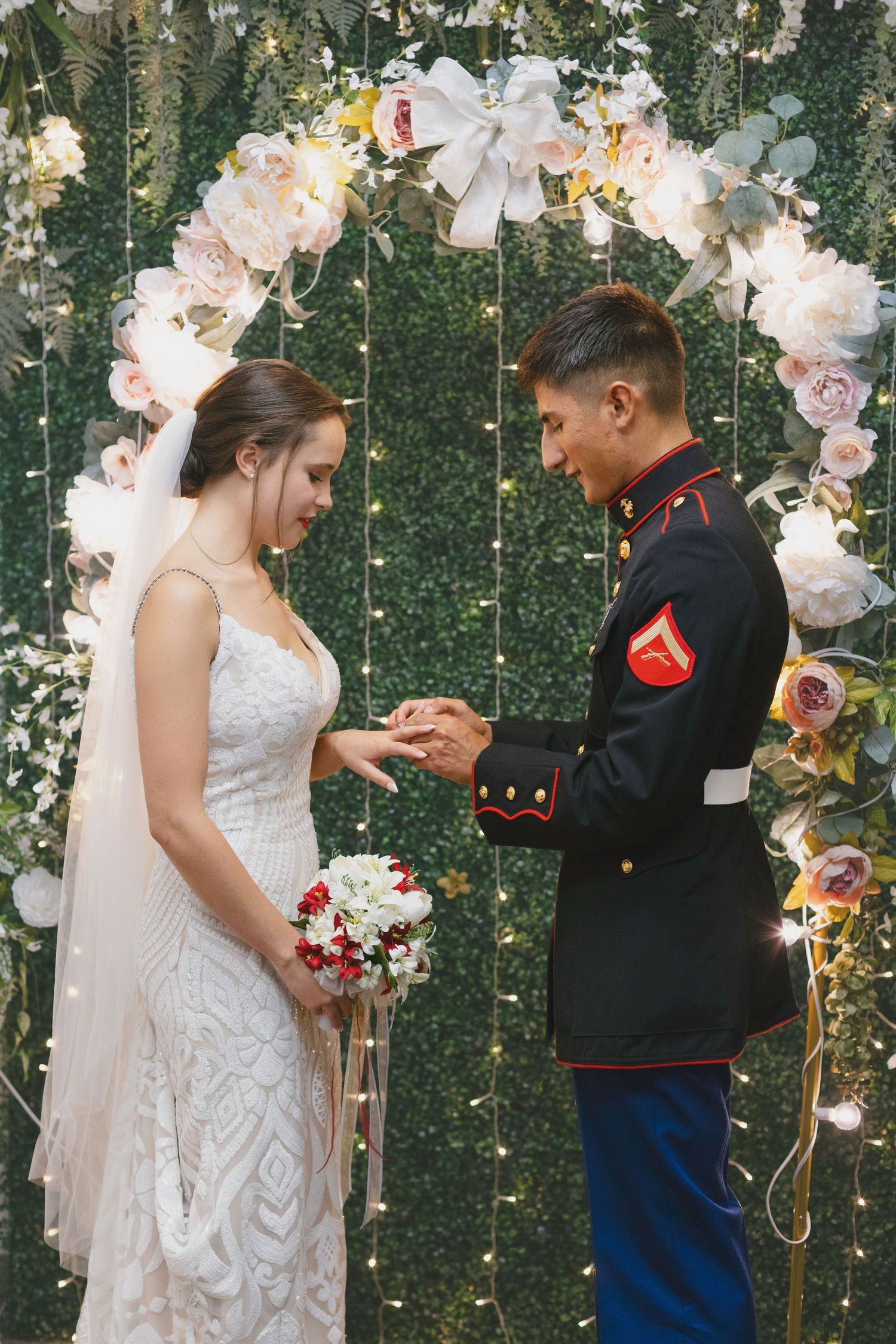 Marine groom places wedding ring on bride's finger under floral arch with fairy lights.