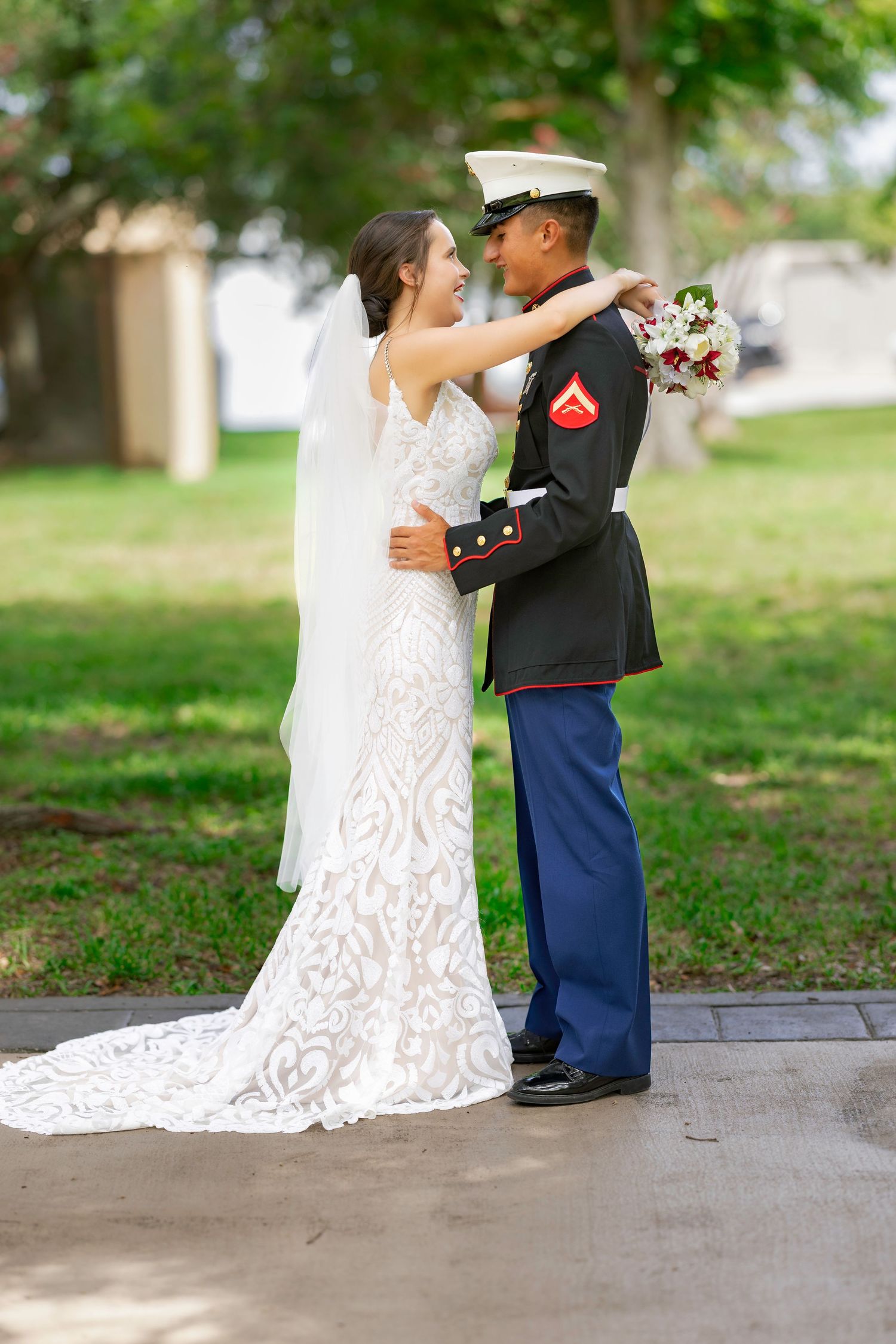 A bride in lace gown and Marine groom in dress blues embrace outdoors on their wedding day.
