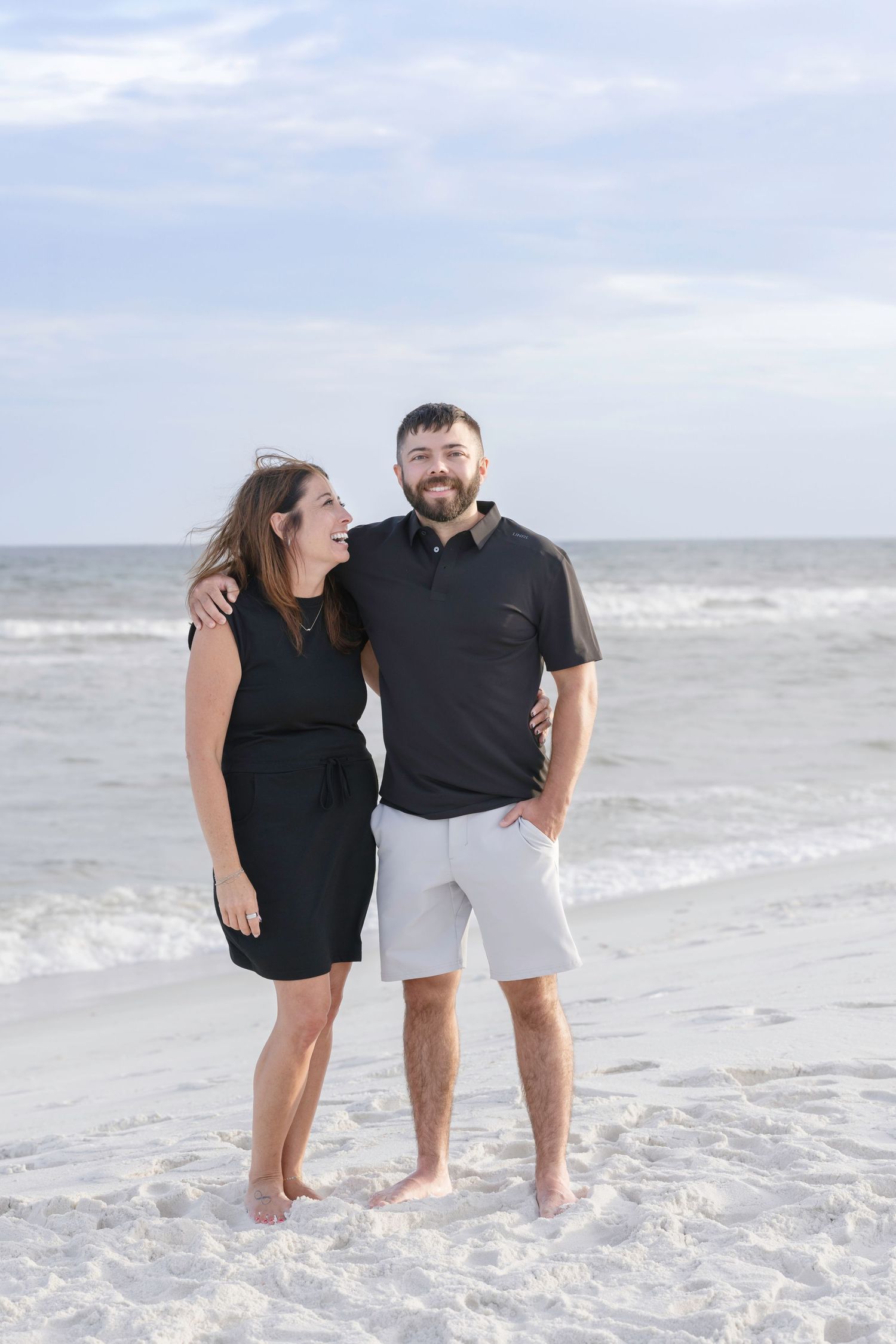 Couple in black outfits smiling together on a sandy beach with ocean waves in the background.