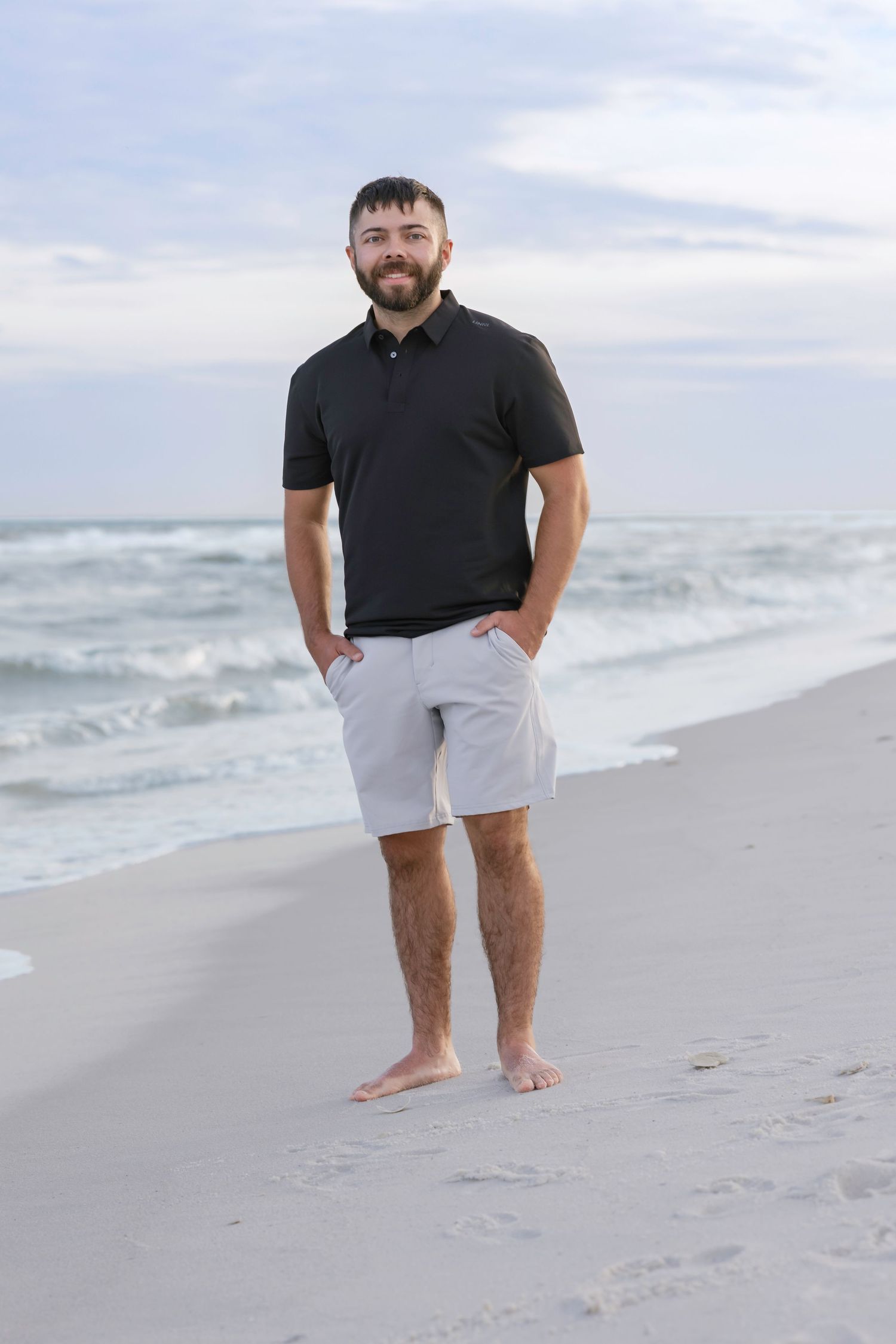 A smiling bearded man in a black polo shirt and gray shorts stands barefoot on a sandy beach with ocean waves behind him.