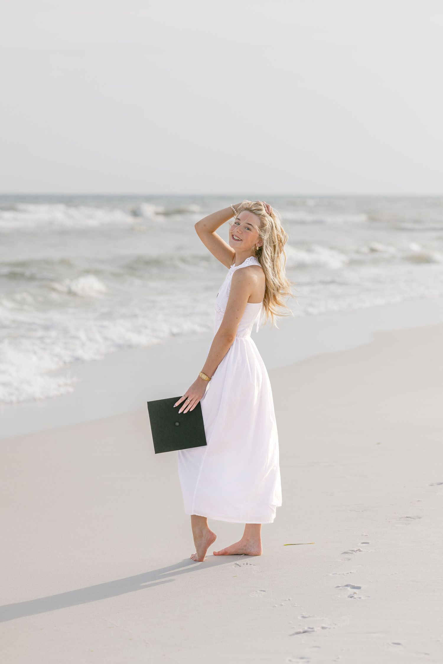 Smiling blonde woman in white dress holding graduation cap on sandy beach with ocean waves.