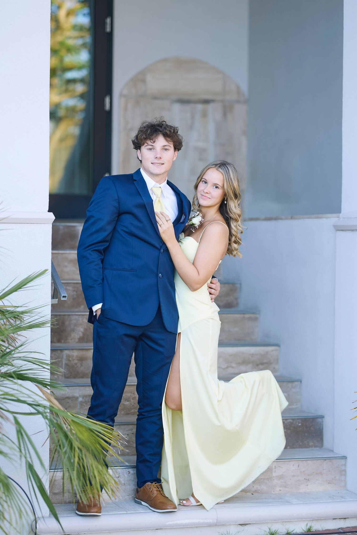 A couple in formal attire poses playfully on white stone steps with palm trees in the background.