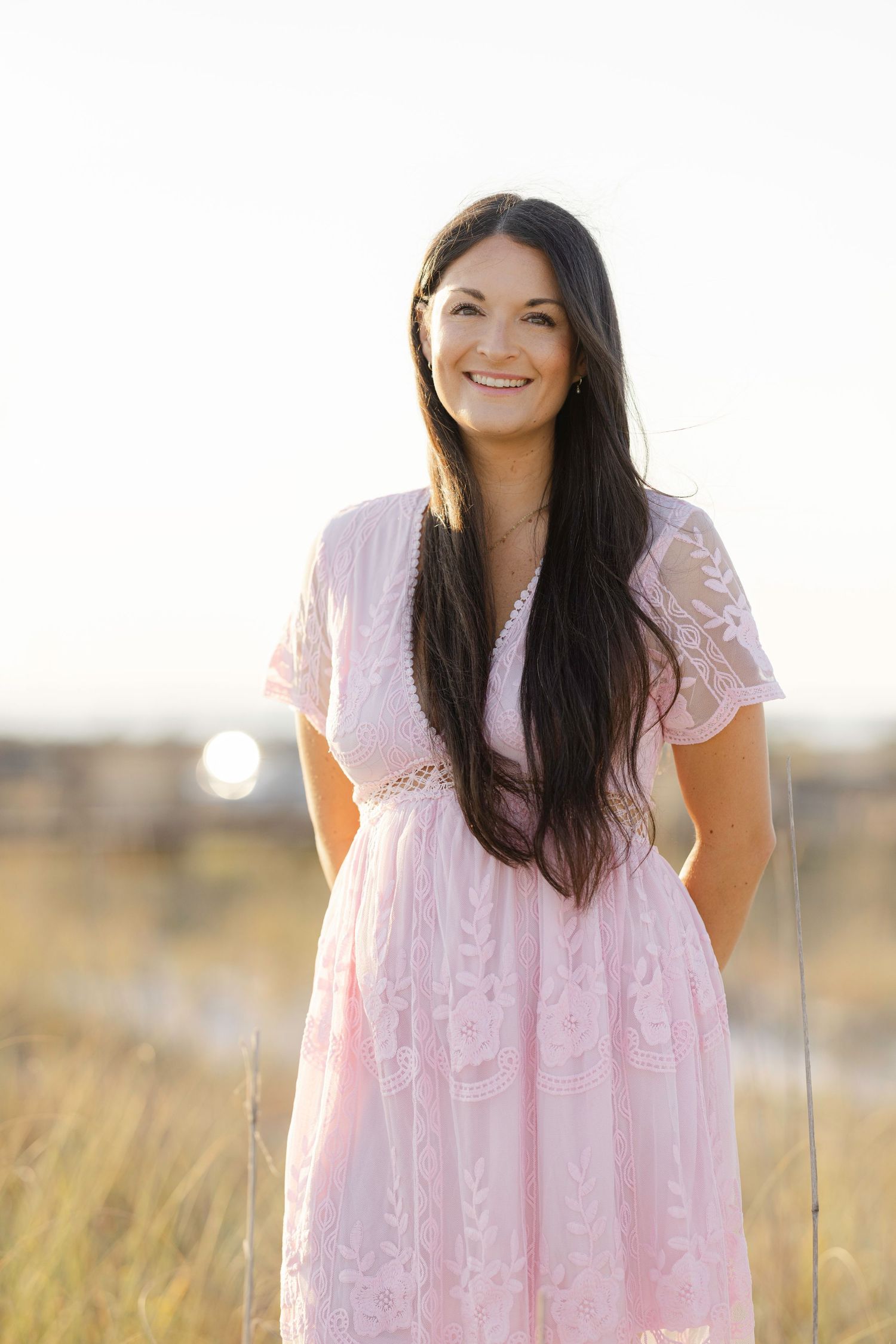Smiling young woman with long dark hair wearing a pink lace dress standing in a golden field at sunset.